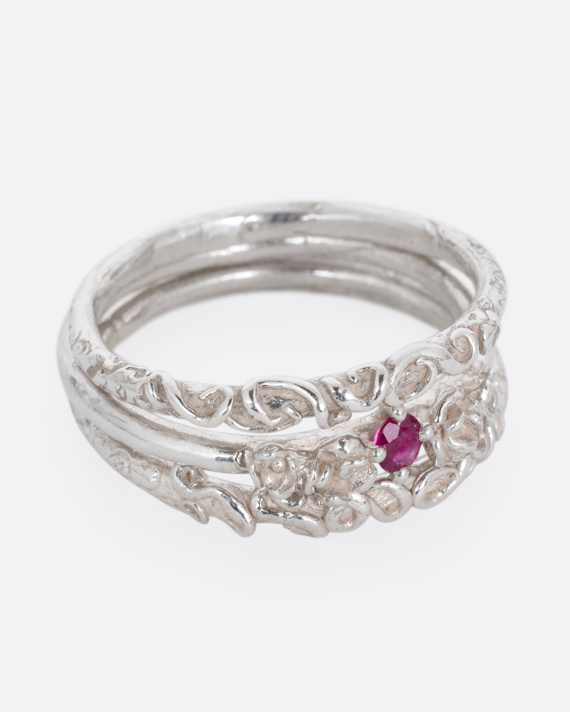 A silver ring with three bands and a round prong set ruby at the center, shown from the side.