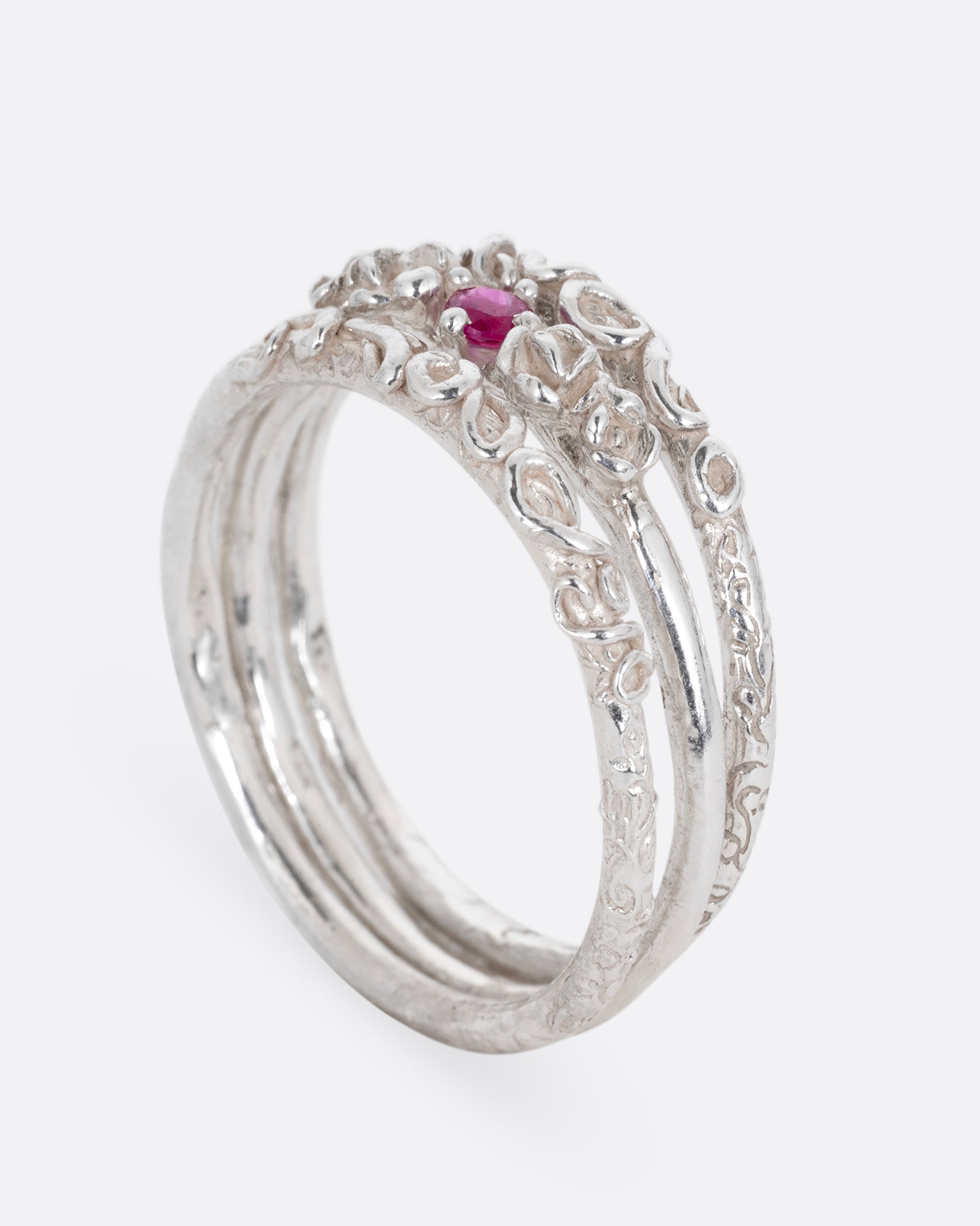 A silver ring with three bands and a round prong set ruby at the center, shown standing up from the side.