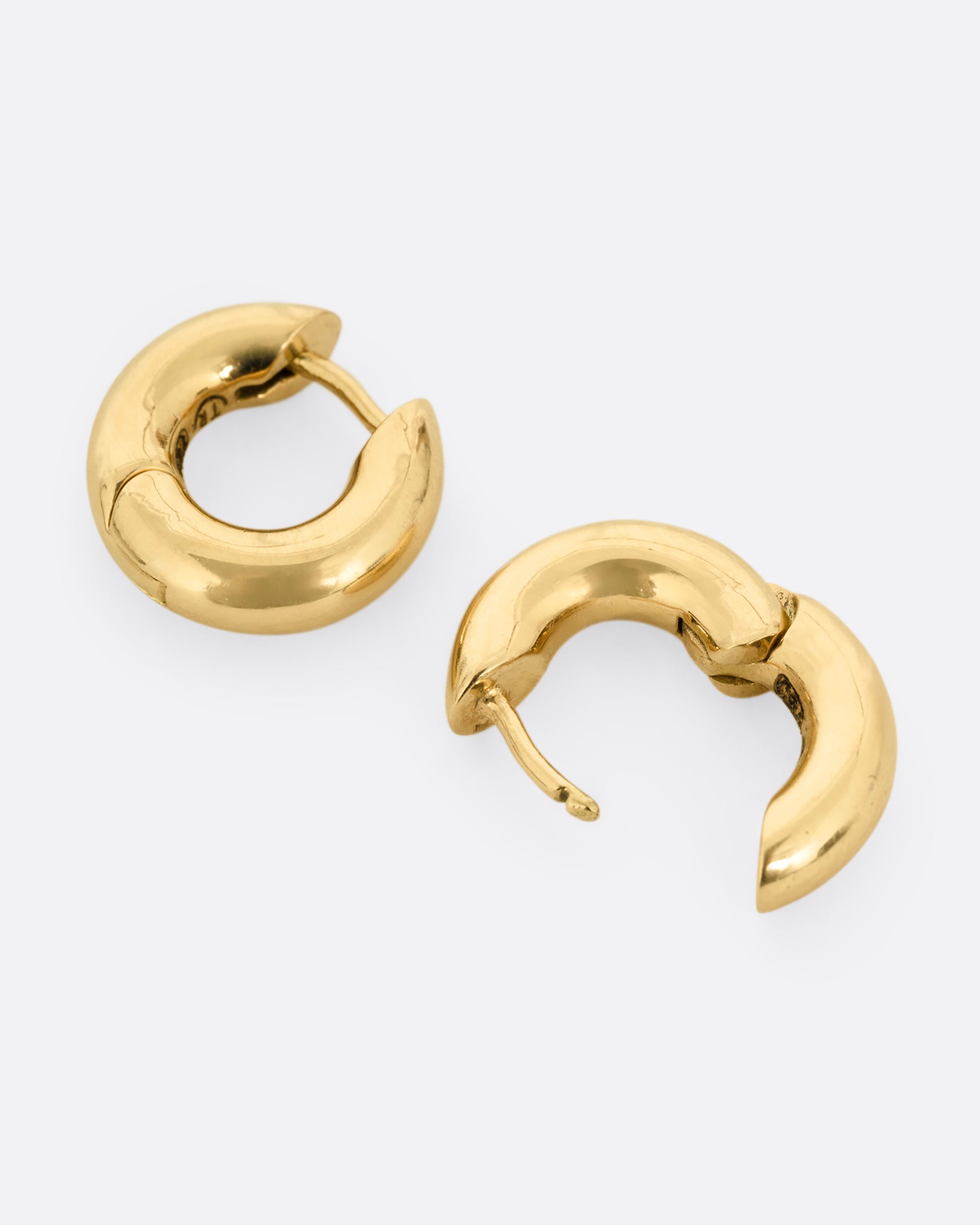 A pair of yellow gold huggie hoops with hinges and clicker clasps, shown lying flat from the side. One earring is shown open.