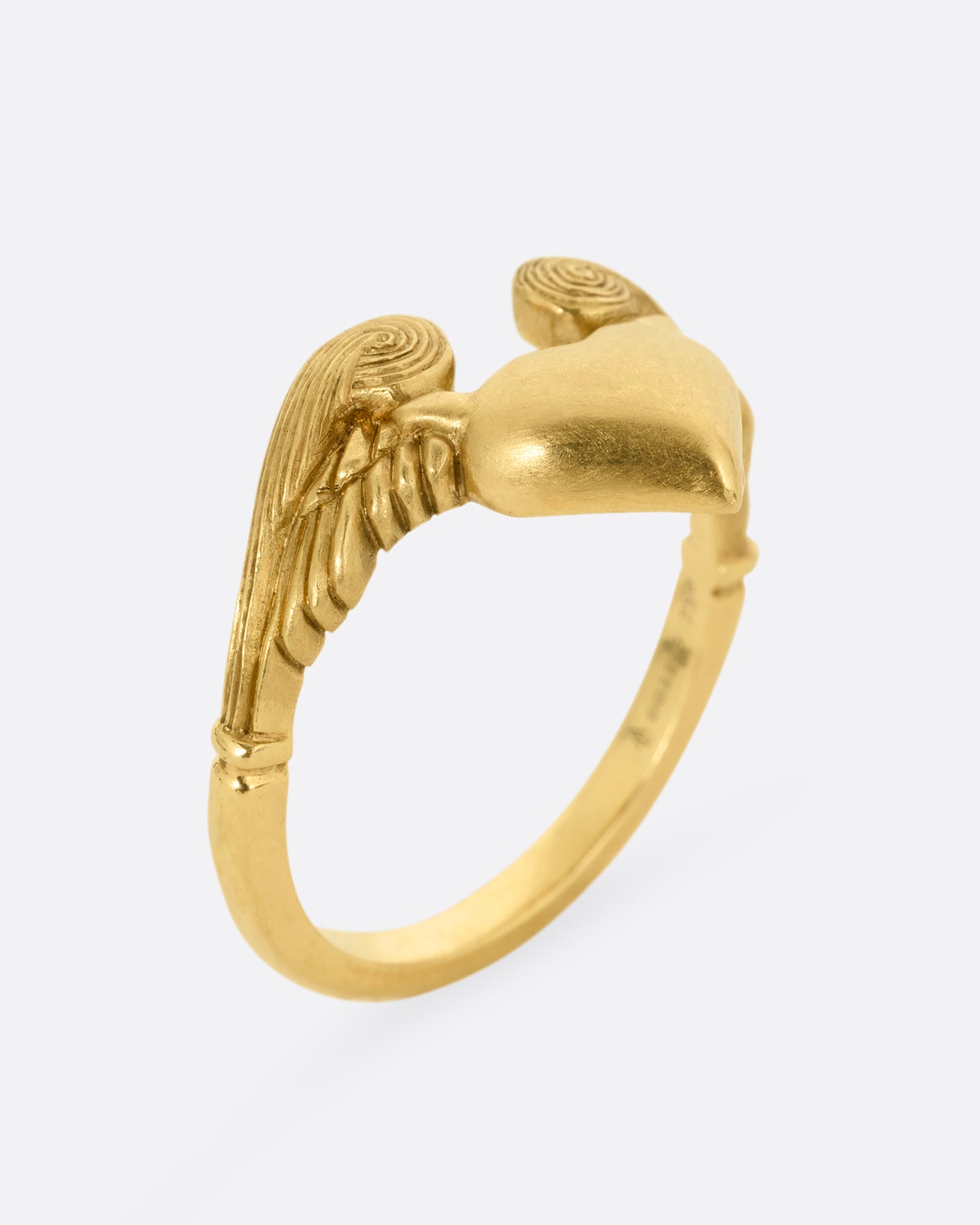 A yellow gold ring with a heart with wings on it. Shown from the side, standing up.