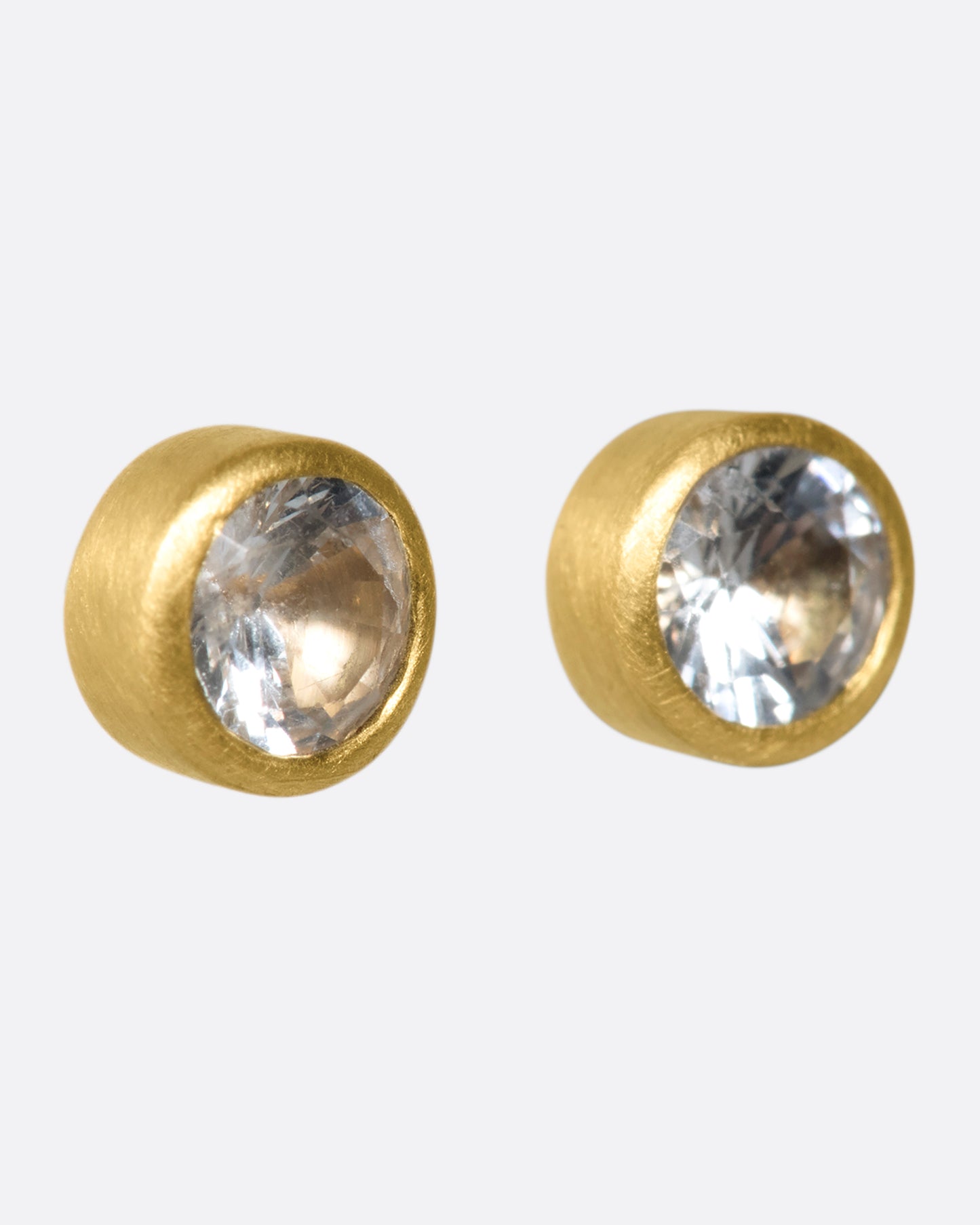 A pair of yellow gold solitaire earrings with round white sapphires in bezel settings.