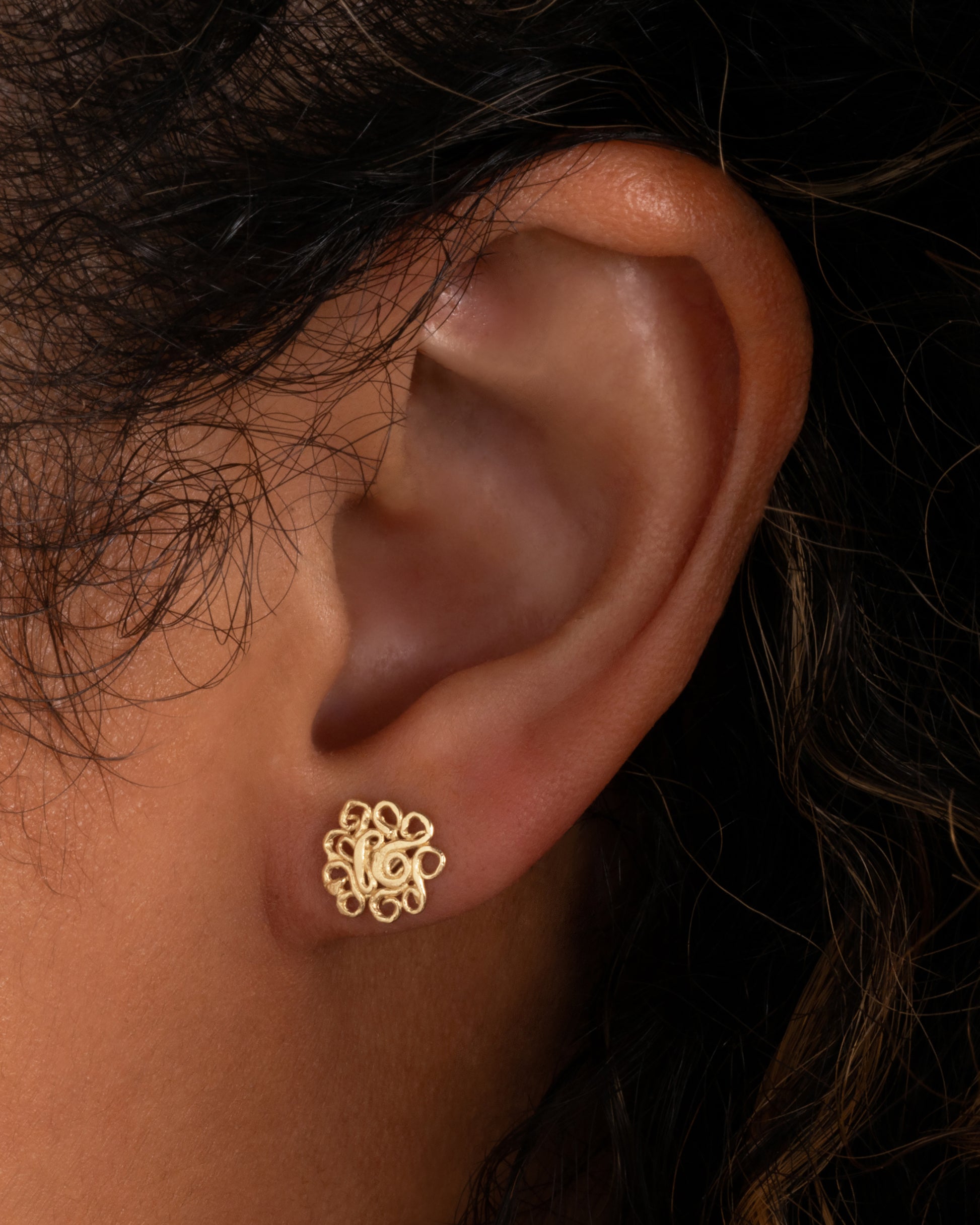 A pair of yellow gold stud earrings with swirling vines in a lacy pattern. Shown worn in an ear.