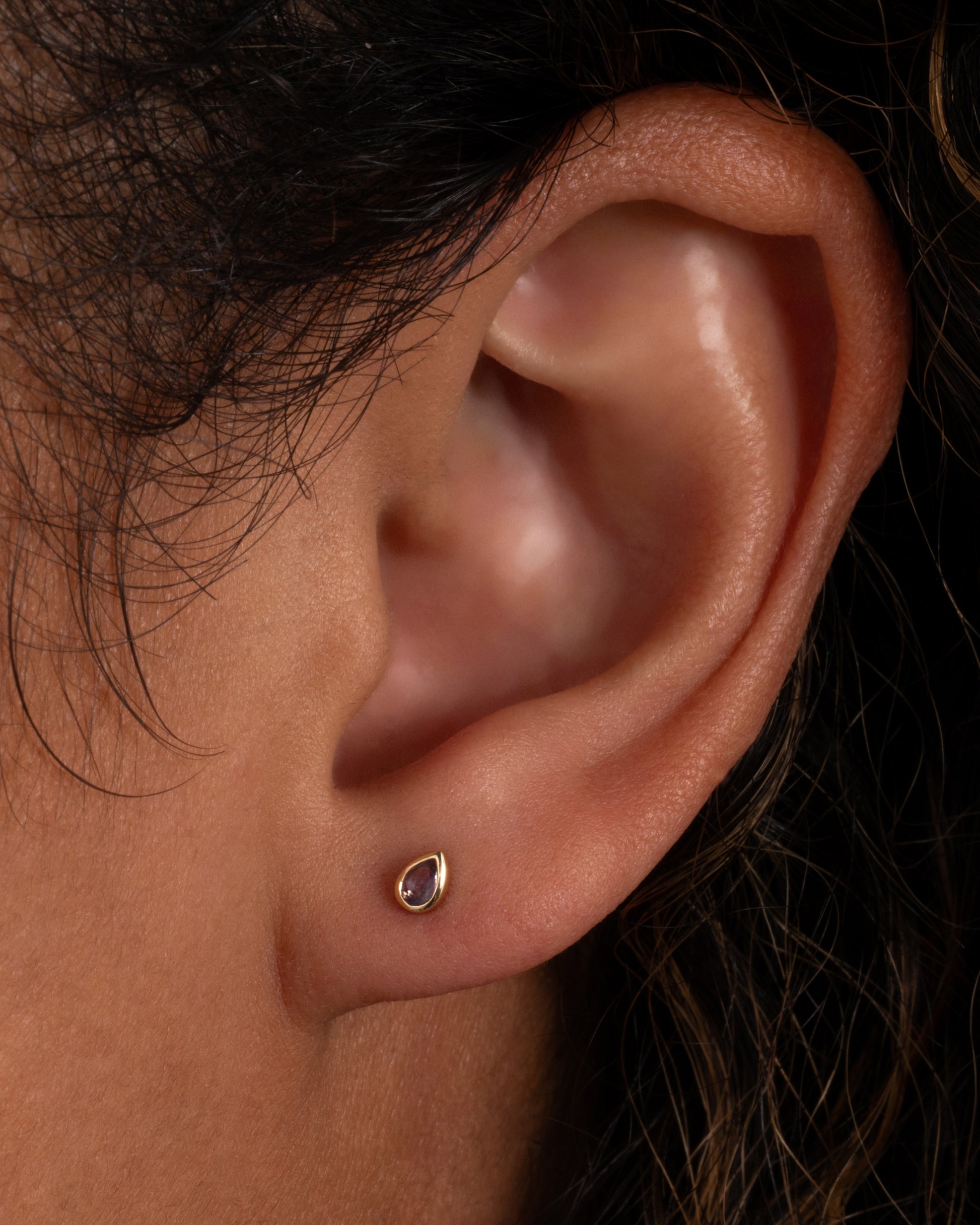 A yellow gold stud earring with a pear shaped lavender shone, shown on an ear.
