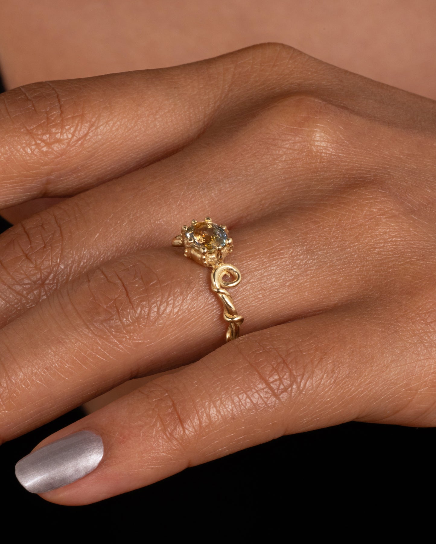 A twisty yellow gold ring with a bicolor sapphire, shown worn on finger.