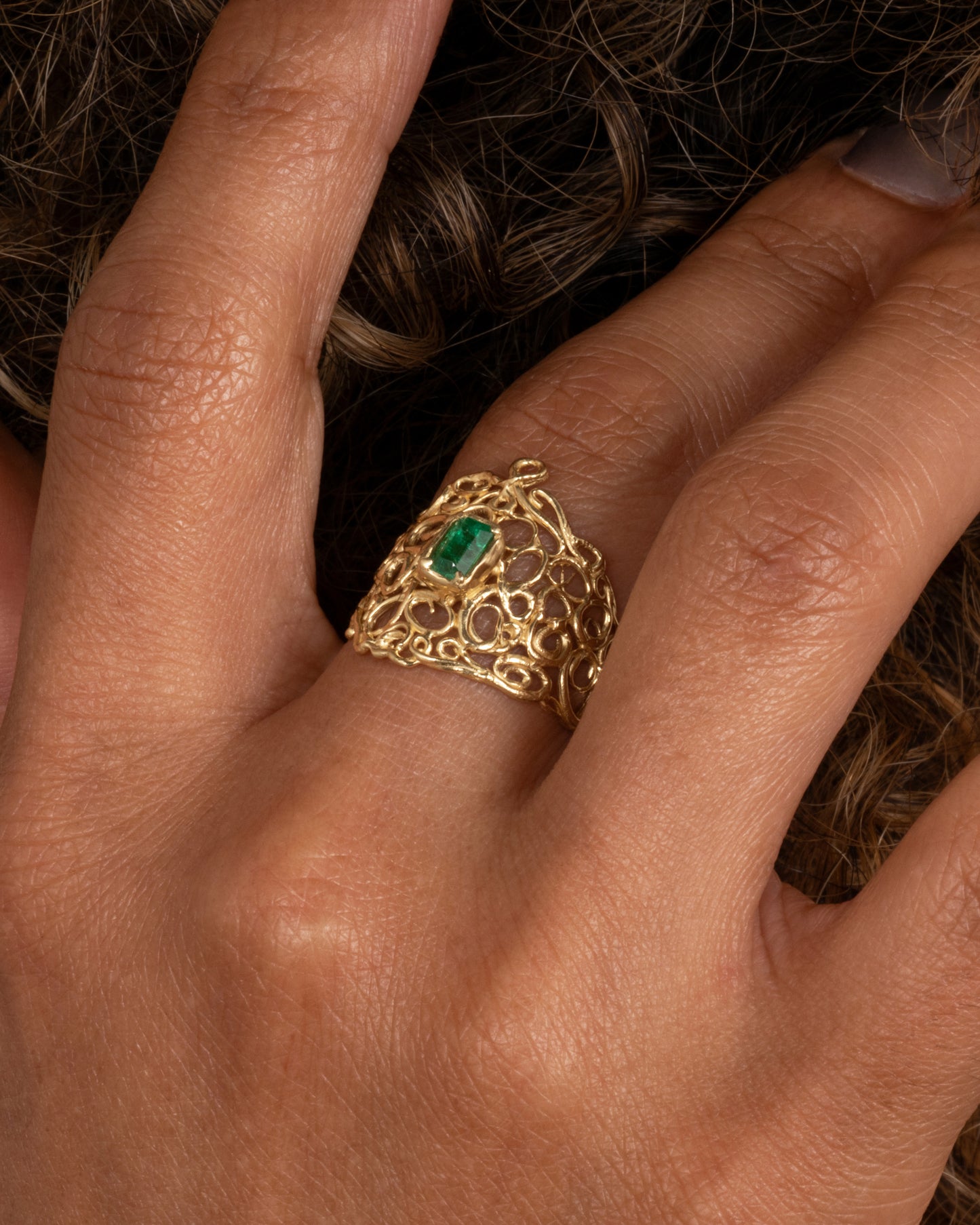 A wide yellow gold curling vine ring with a central emerald. Shown worn on finger.