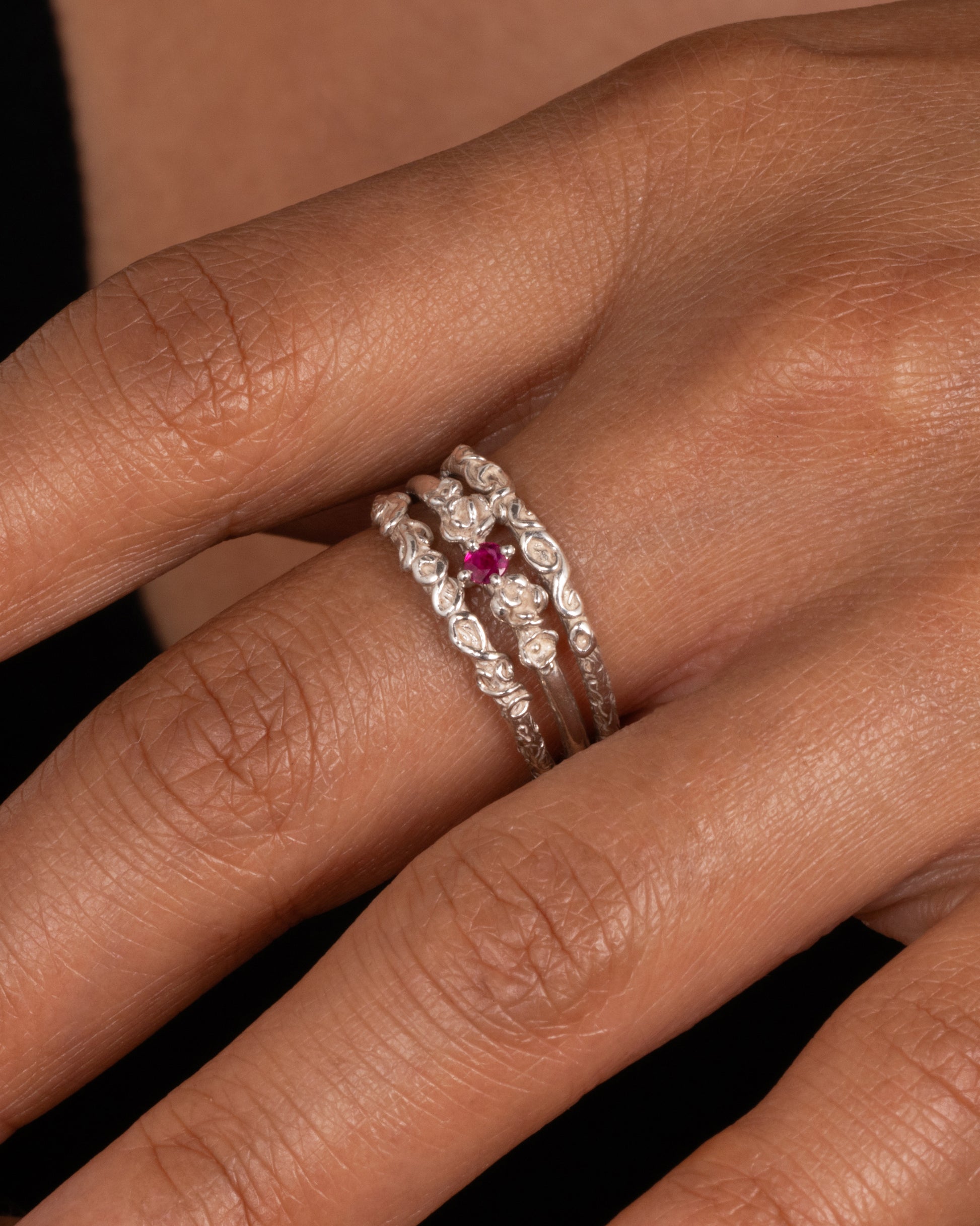 A silver ring with three bands and a round prong set ruby at the center, shown worn on finger.