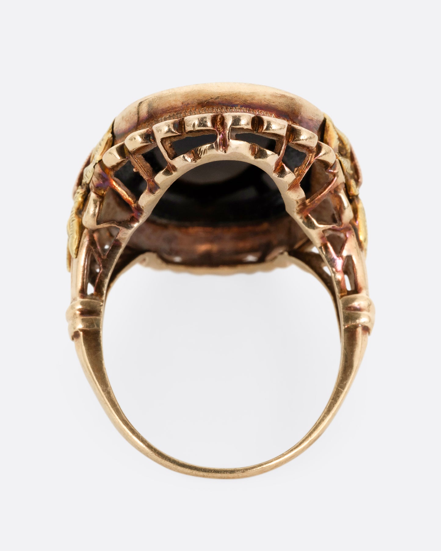 A yellow, rose, and green gold ring with a scalloped bezel, bow- topped shoulders, and an oval onyx. Shown from the back.