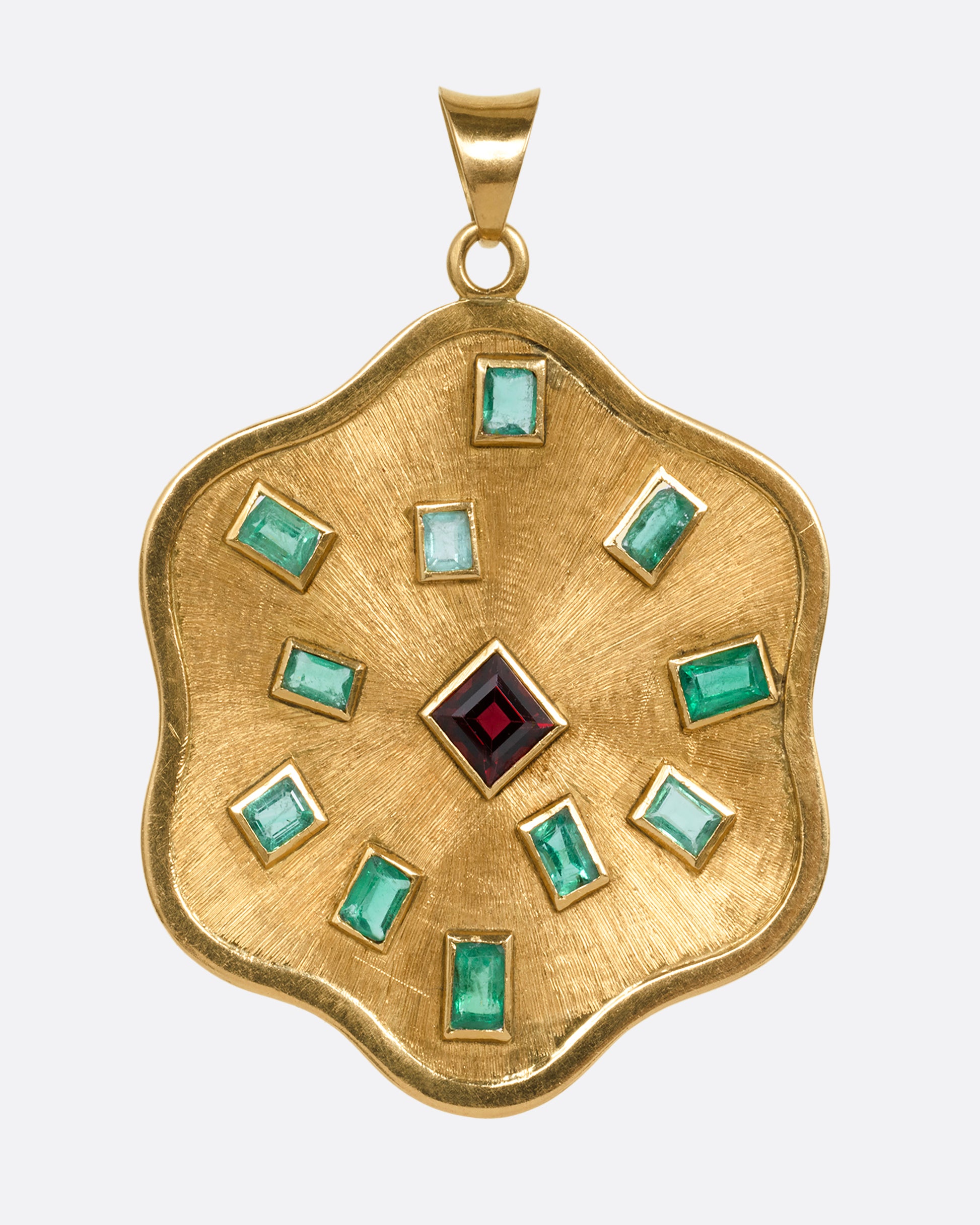 A yellow gold wavy edged medallion pendant with baguette emeralds encircling a central princess cut garnet. Shown from the front.