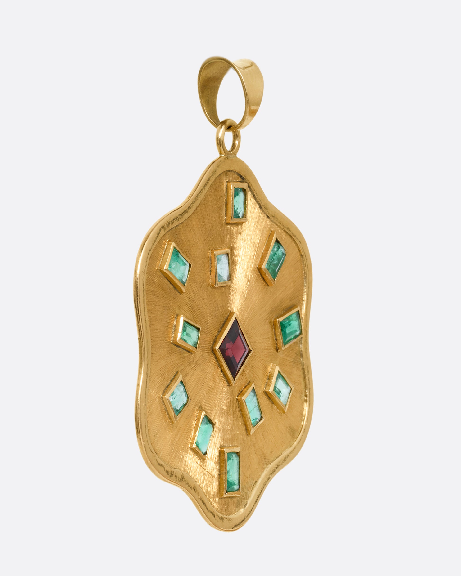A yellow gold wavy edged medallion pendant with baguette emeralds encircling a central princess cut garnet. Shown from the side.