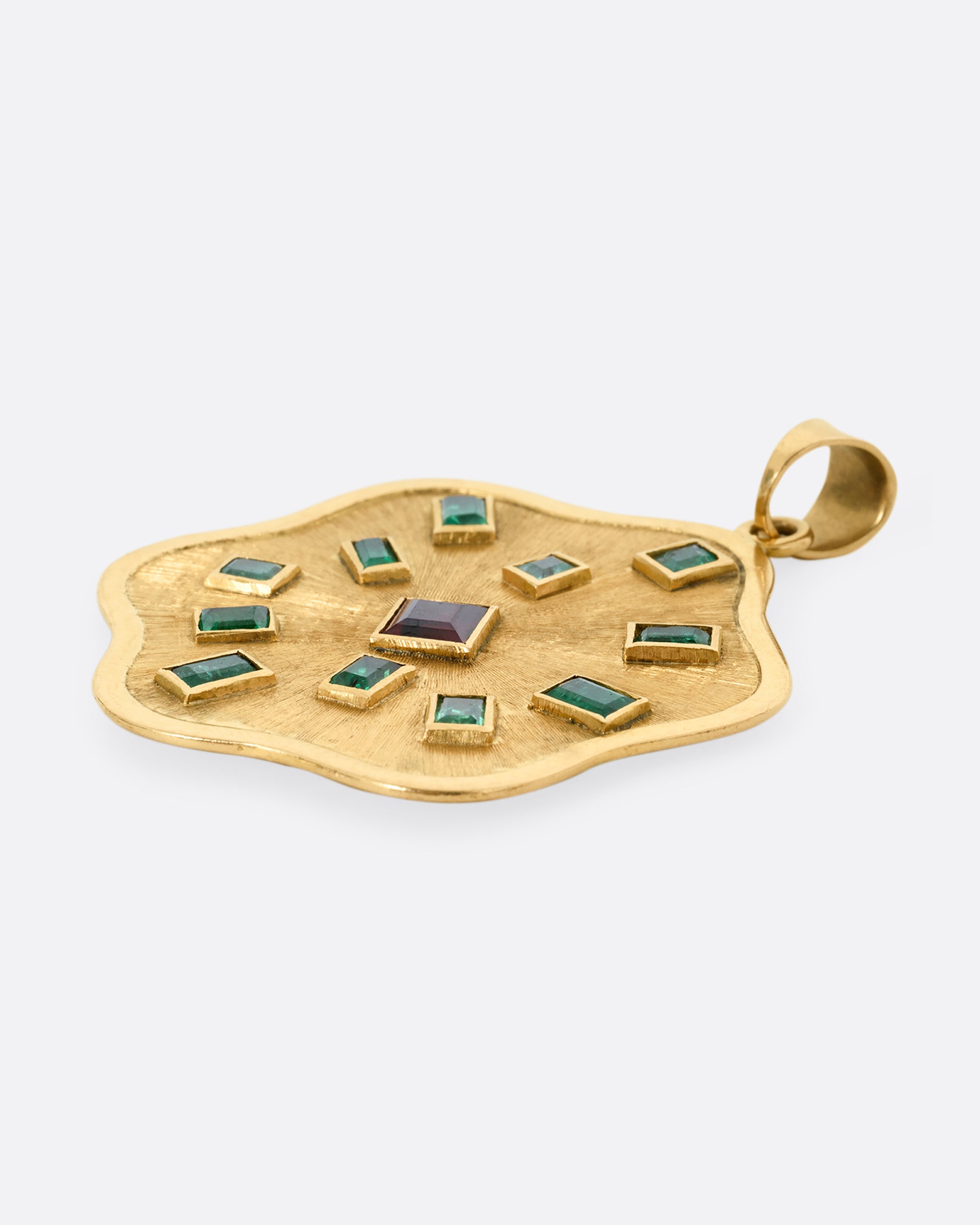 A yellow gold wavy edged medallion pendant with baguette emeralds encircling a central princess cut garnet. Shown lying flat from the side.