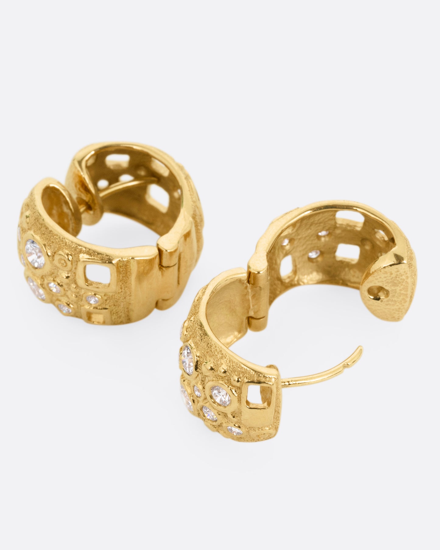 A pair of wide yellow gold huggie hoops with diamonds of varying sizes and small rectangular cutouts, shown lying on their sides with one hoop open.