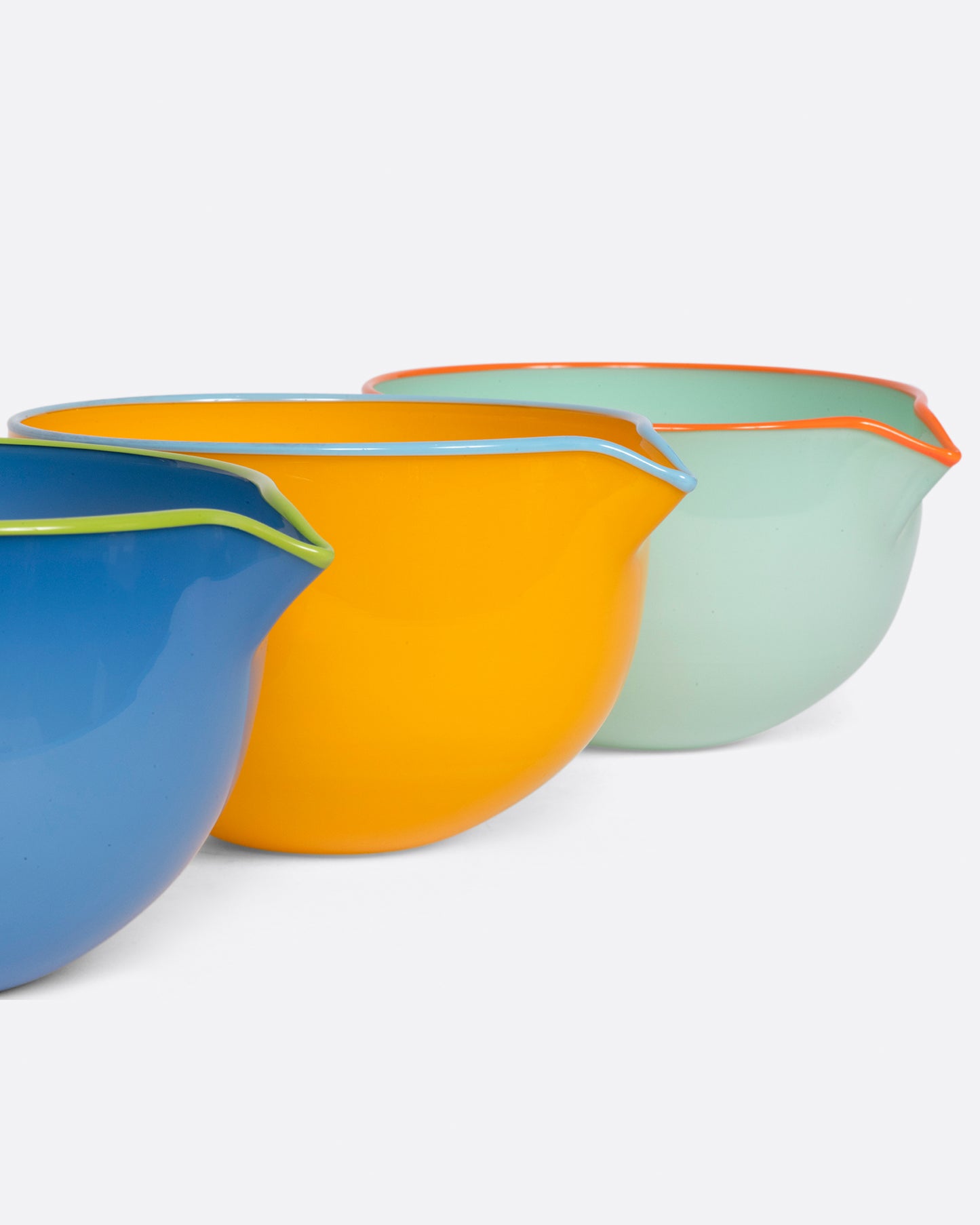A set of three hand blown glass spouted pouring bowls in blue with a chartreuse edge, yellow with a blue edge, and green with an orange edge. Shown from the side arranged in a line.