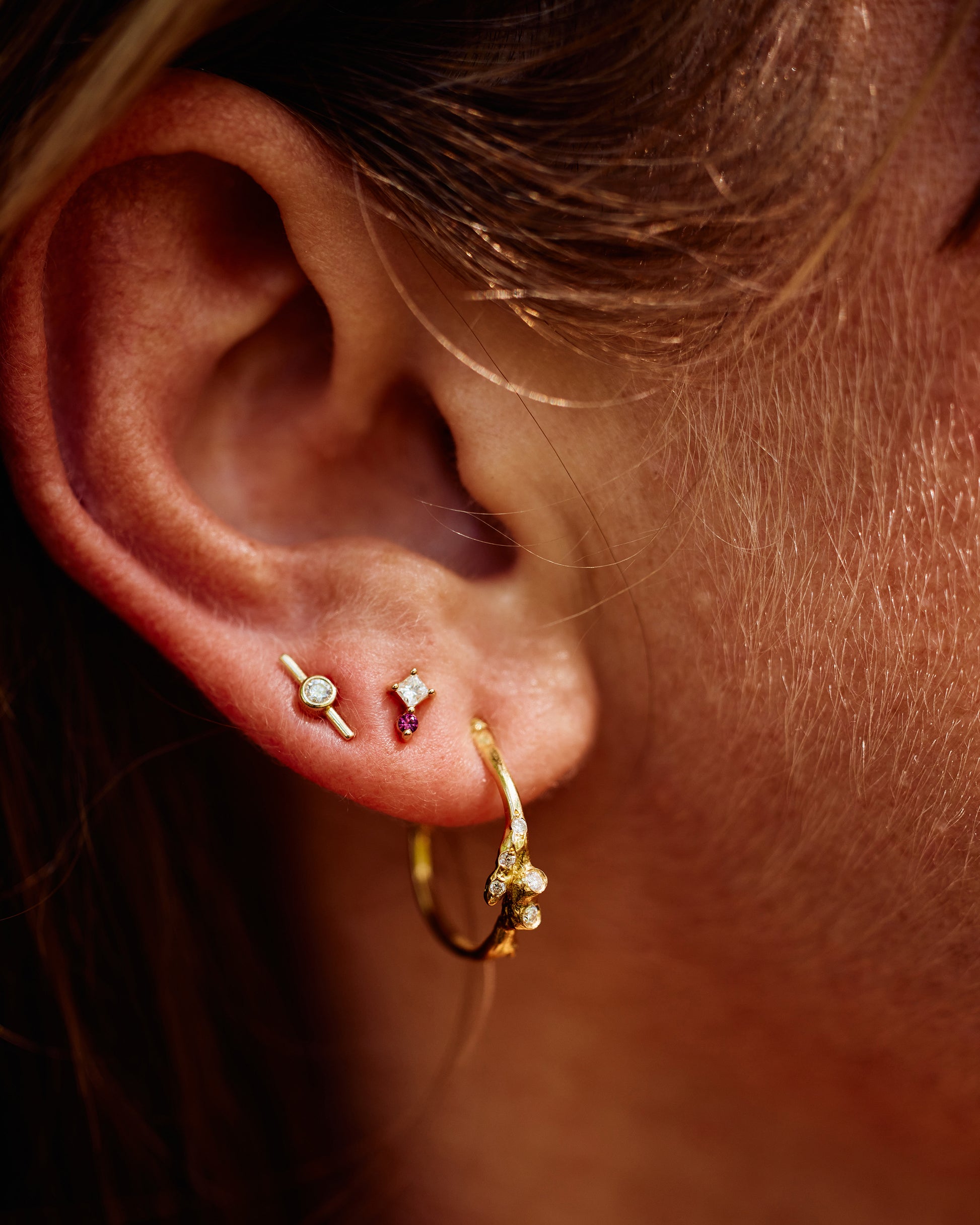 A close up of an ear with three earrings; a yellow gold hoop with diamonds, a stud earring with a princess cut diamond and a round pink sapphire, and a yellow gold diamond stud earring. View from the front.