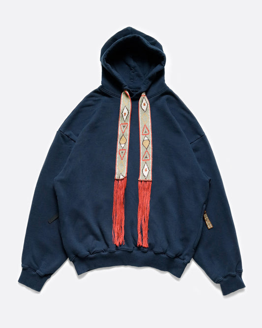 A blue cotton fleece hoodie with an oversized silhouette, accented with wide, fixed drawstrings in a geometric pattern with long red fringe.