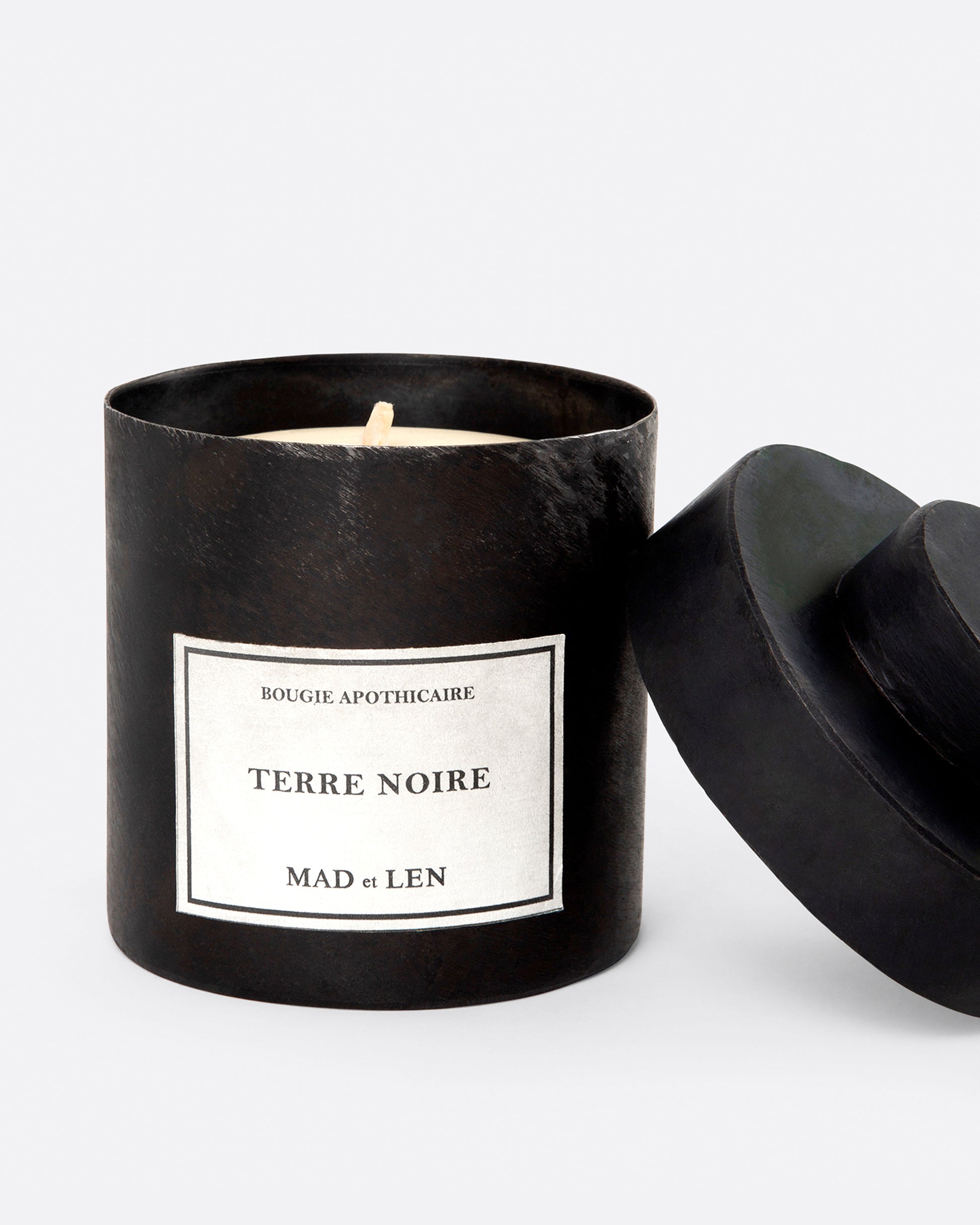 A Mad et Len Terre Noire candle shown with its lid leaning against the side, shown from the front.
