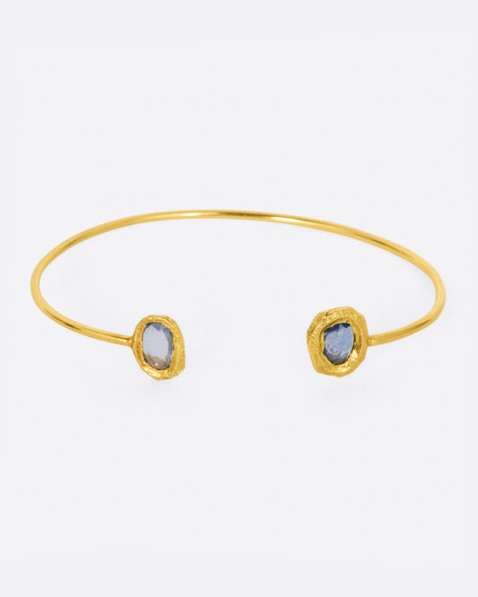 A simple yellow gold wire cuff bracelet with a blue sapphire in a hand carved bezel on either end.