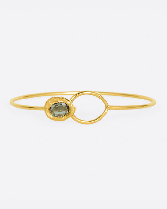 A yellow gold wire bracelet with an open loop at one end and a hook, punctuated by a bezel set green sapphire, on the other.