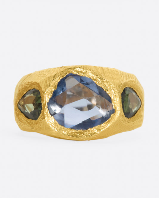 A brushed texture yellow gold ring with a large rose cut blue sapphire at its center with one green pear shaped sapphire on either side.