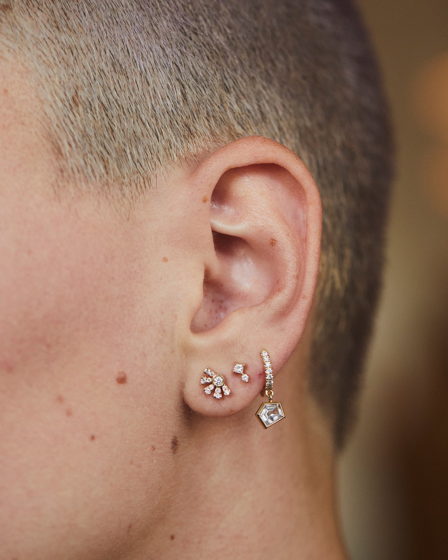 An ear with three diamond earrings. The highest is a hoop with a dangle. The middle is two diamonds stacked. The lowest is sun shaped. Shown from the front.