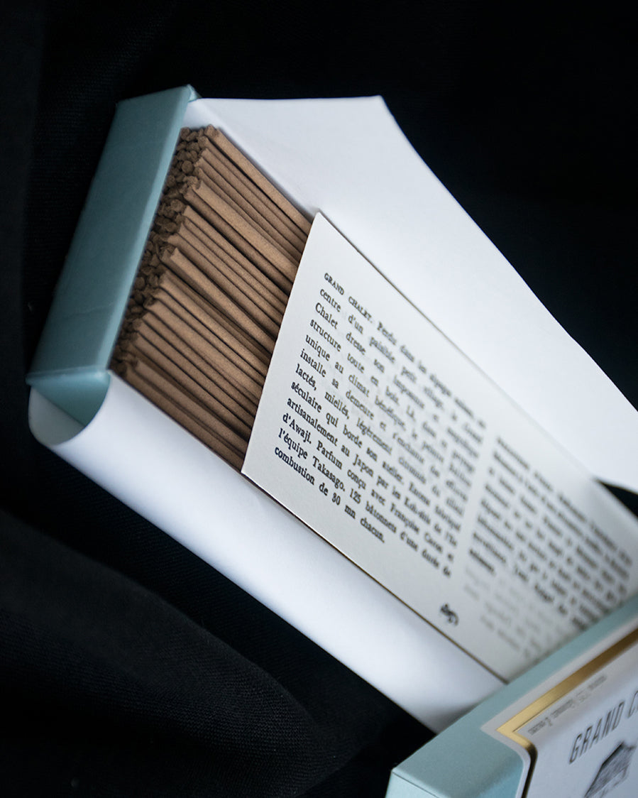 An open box of incense revealing incense sticks and the included literature as well as the turquoise, white, and metallic gold packaging. Shown diagonally from the top.