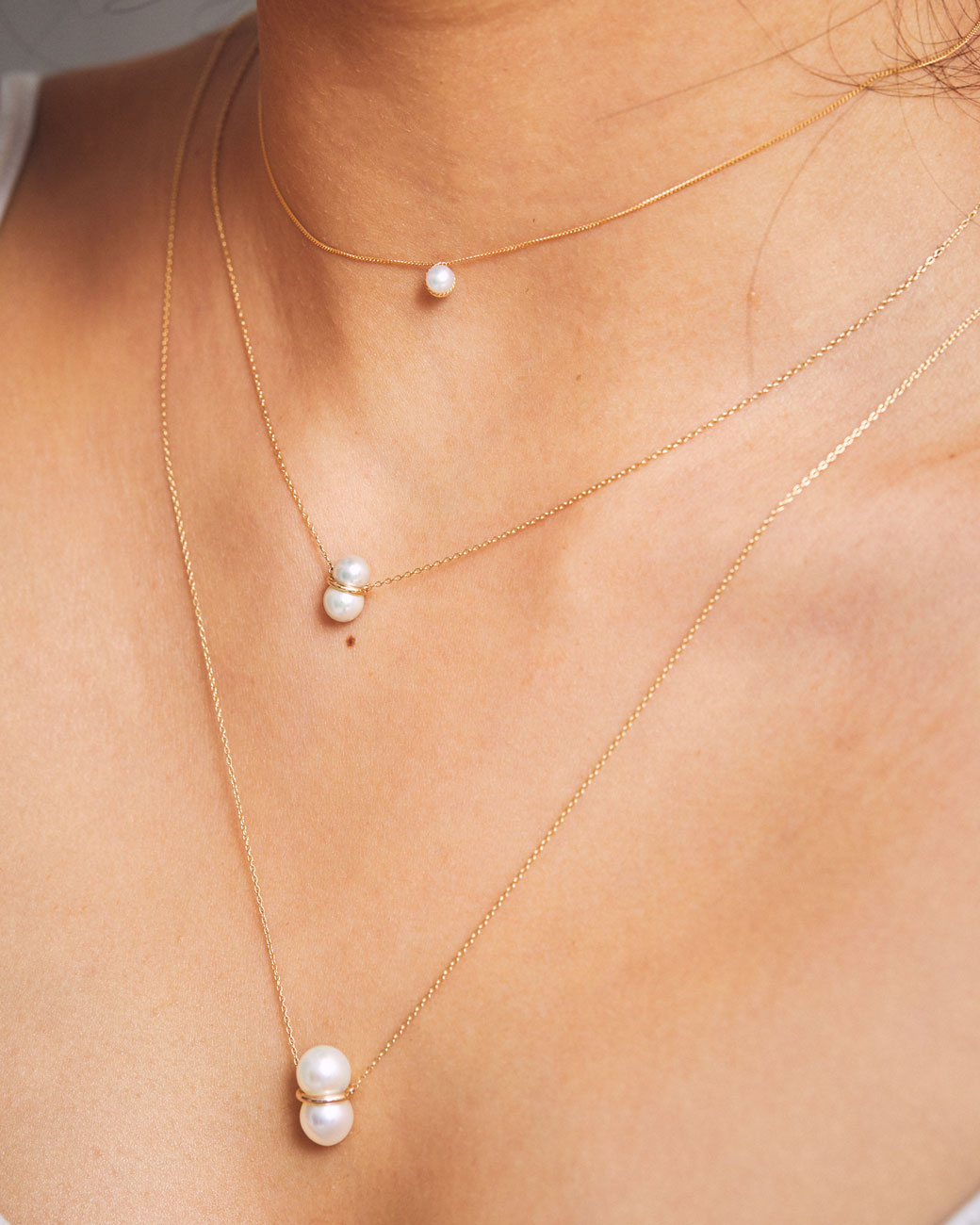 A woman's neck with three pearl necklaces of different sizes on. The shortest is a single pearl, the middle is a small twin pearl, and the longest is a large twin pearl. Shown from the front.