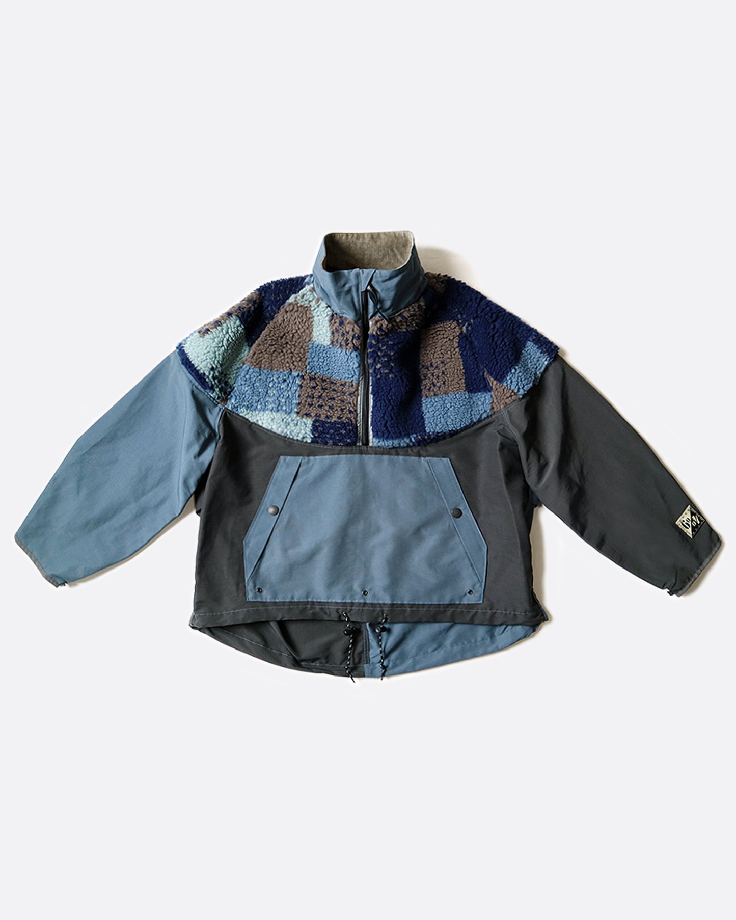 A pullover anorak with a half zip. Half blue and half gray with patchwork fleece around the shoulders. Shown laying flat from the front.
