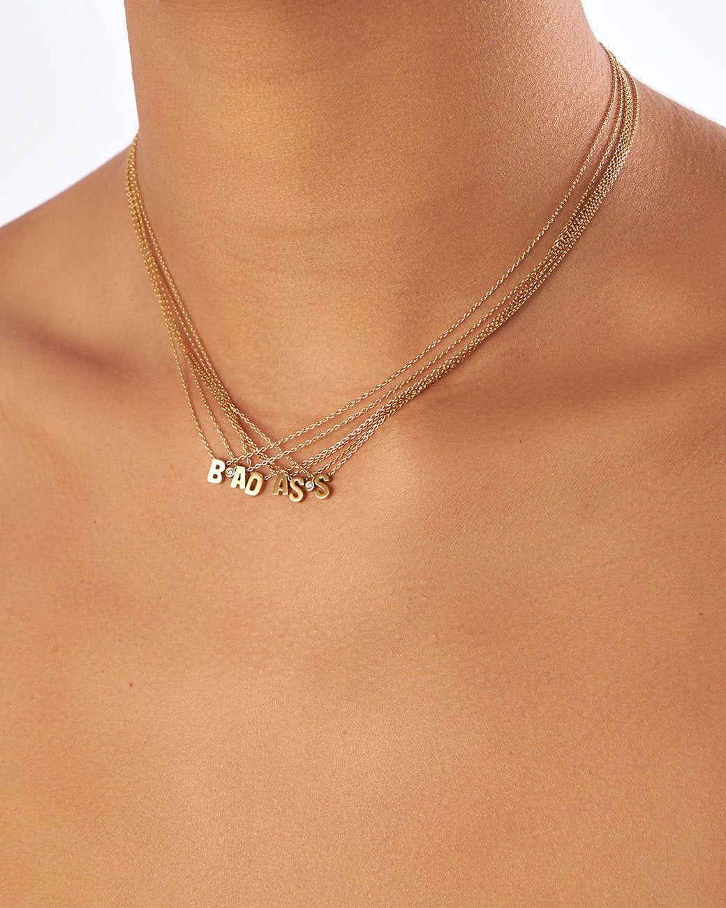 Six yellow gold cable chain necklaces with initial pendants. Shown layered with diamond tags to spell 'BAD ASS' from the front on a person.