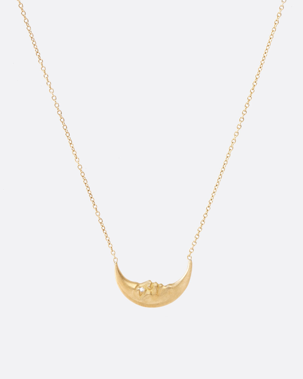 Close up front view of a tiny yellow gold crescent moon, with a face carved into it and little diamond eyes, is a unique pendant. Double sided and hanging from a chain. The chain is attached to the ends of the crescent, so it falls on a sideways/horizontal angle across the chest.