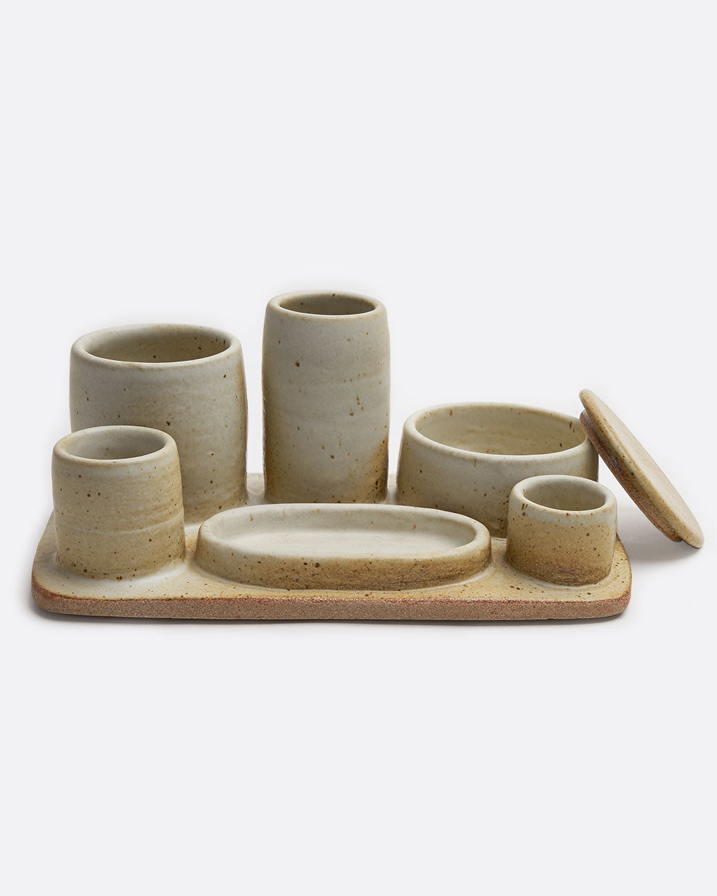 A stoneware pottery organizer tray with various sized cups, jars, and a dish in a matte beige finish. Shown from the front with the jar lid leaning on one side.