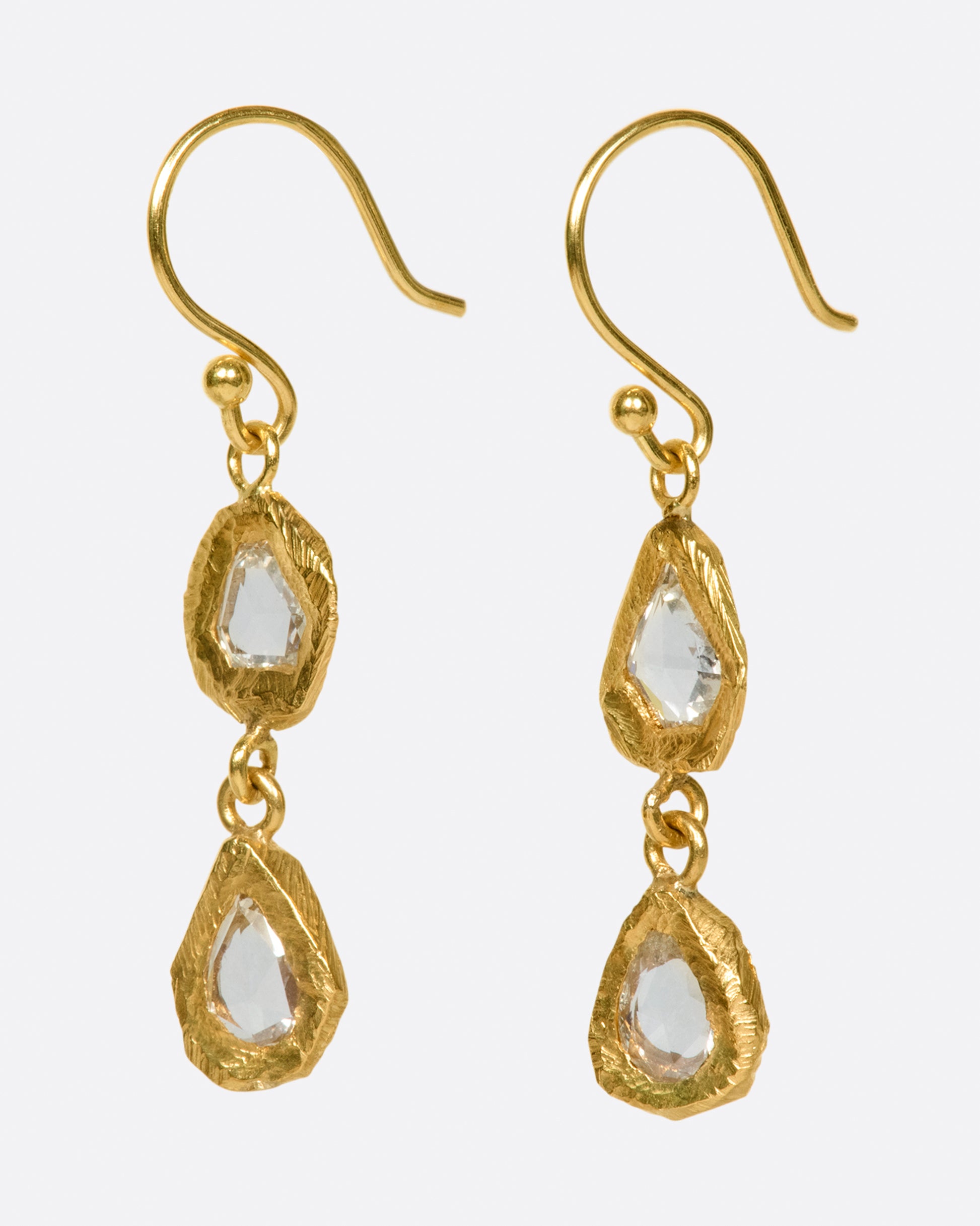 A pair of slightly asymmetrical yellow gold diamond drop earrings with two diamonds set in hand carved bezels on each earring. Shown from the side.