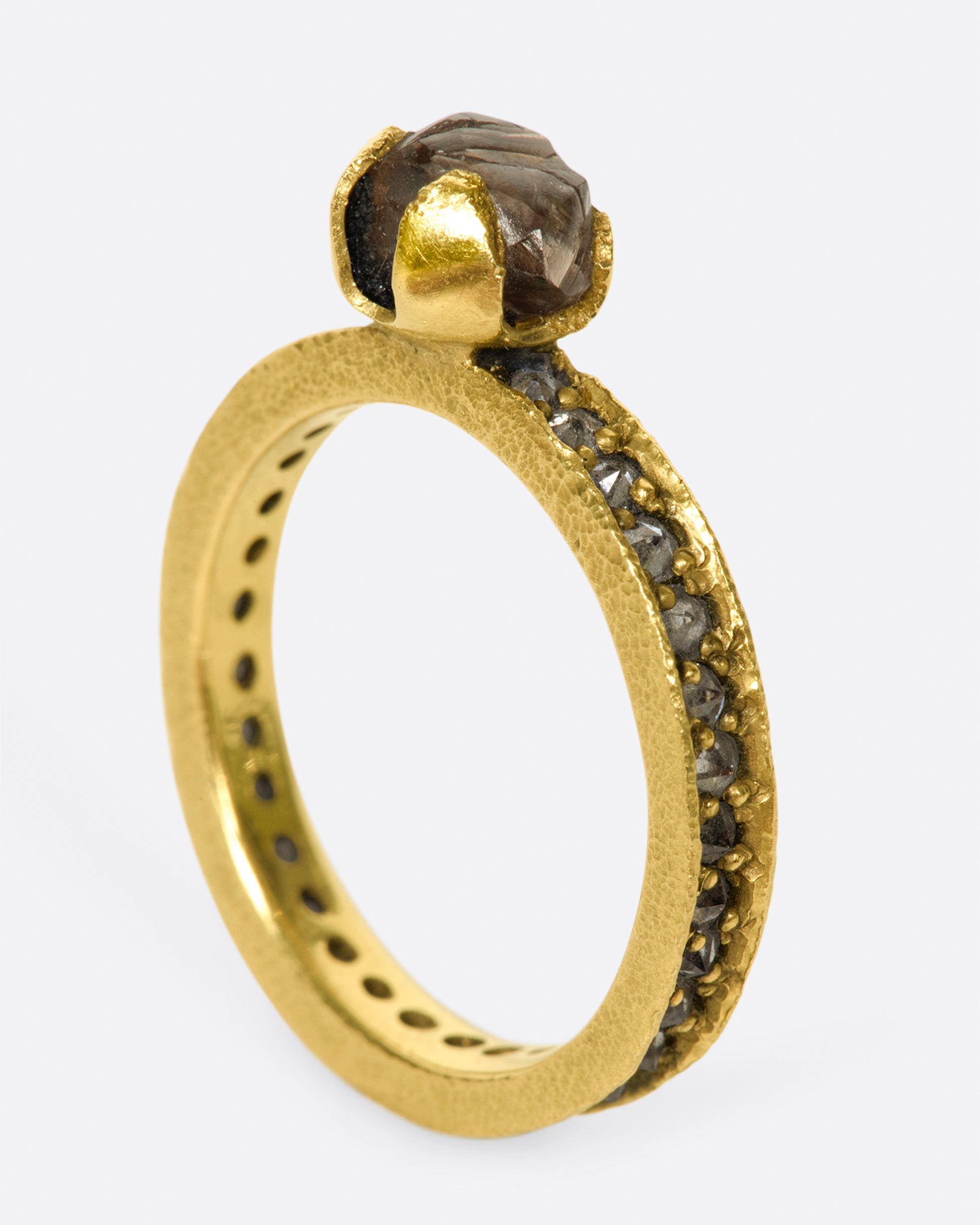 A yellow gold eternity band with inverted diamonds, crowned by a prong set raw yellow diamond. Shown standing up from the side.