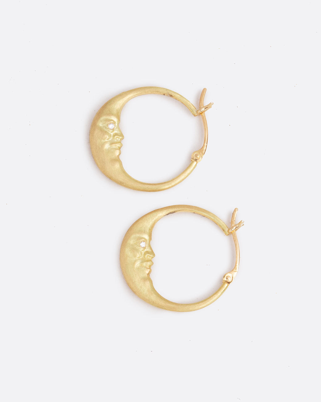 A pair of yellow gold crescent moon hoop earrings with faces and diamond eyes. Each hoop has a clasp at the top. Shown lying flat from the side.