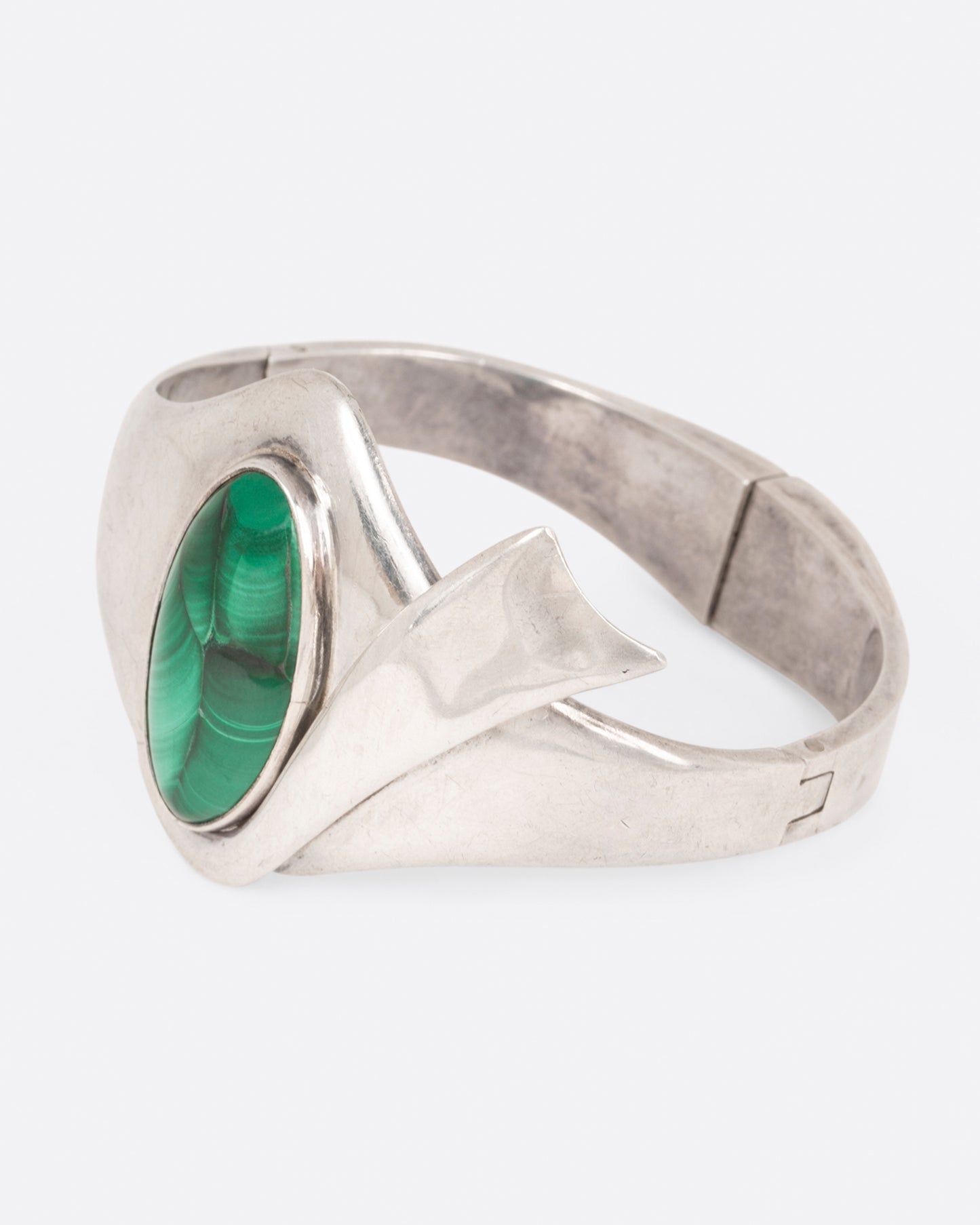 A sterling silver hinged bracelet with a large oval malachite at the center and a ribbon of silver draping off the side, shown from the side.
