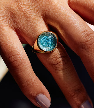 A round aquamarine ring with a sun behind it, worn on the hand.