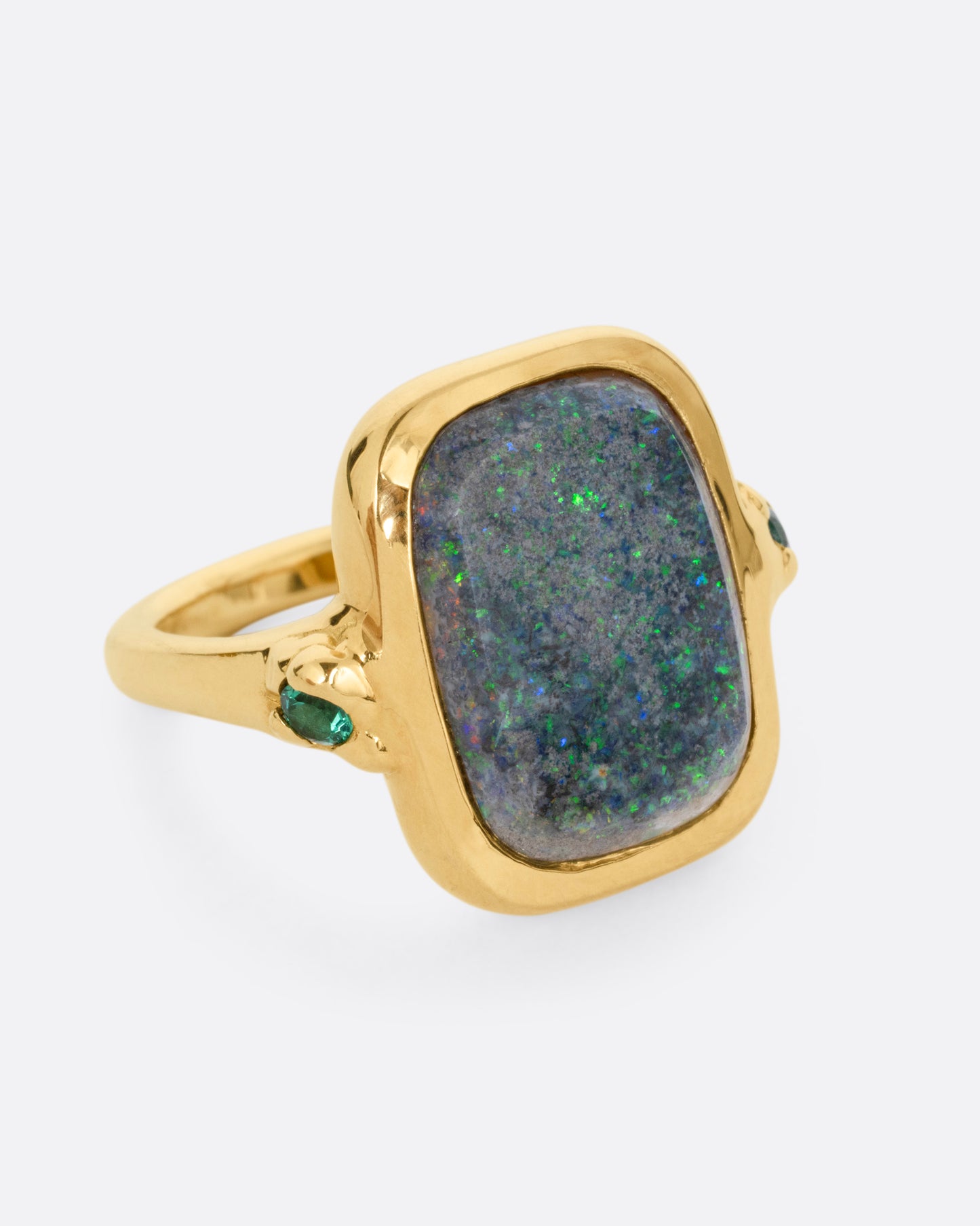 A yellow gold ring with a large rectangular boulder opal and emerald accents, shown from the side.