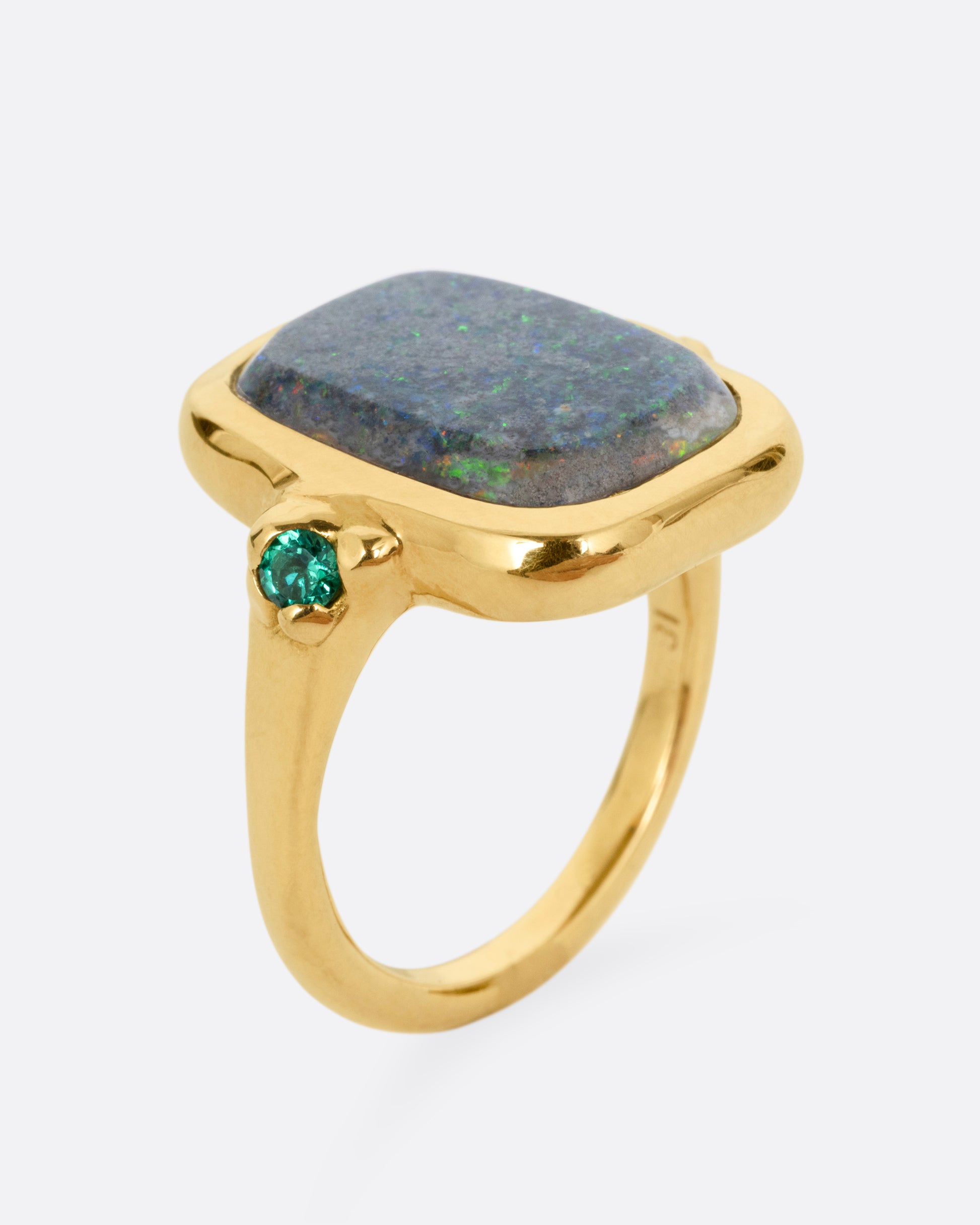 A yellow gold ring with a large rectangular boulder opal and emerald accents, shown from the side standing up.