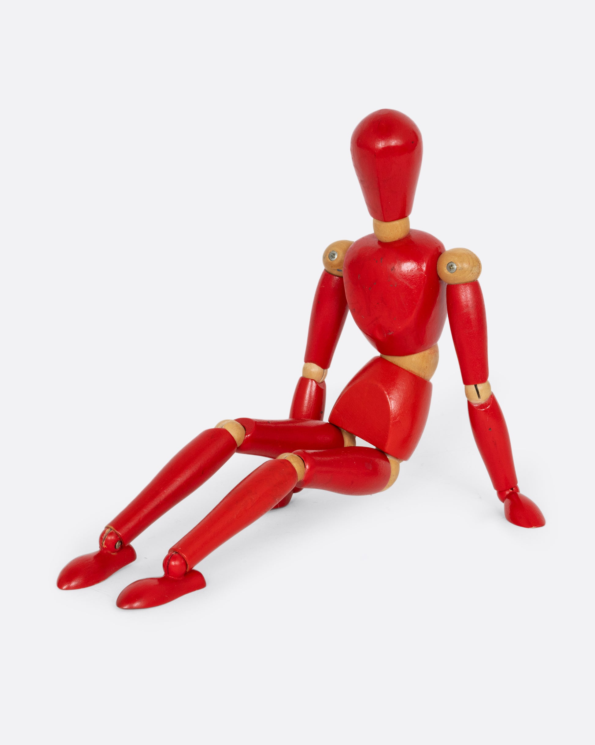 A red wooden articulated human figure. Shown sitting, from the side.