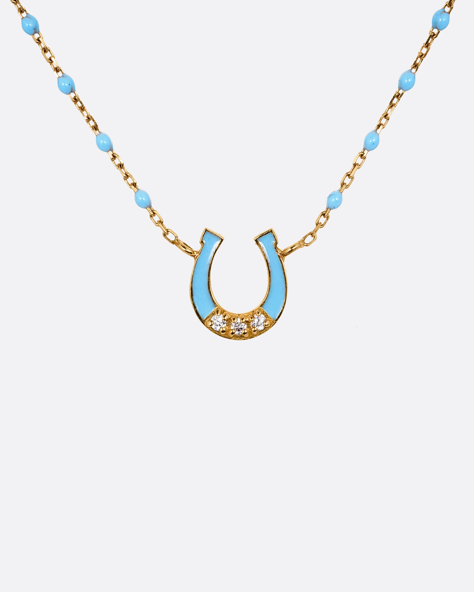 A yellow gold cable chain necklace with droplets of blue resin and a horseshoe charm. The horseshoe charm is gold with blue enamel and three white diamonds. Shown from the front.