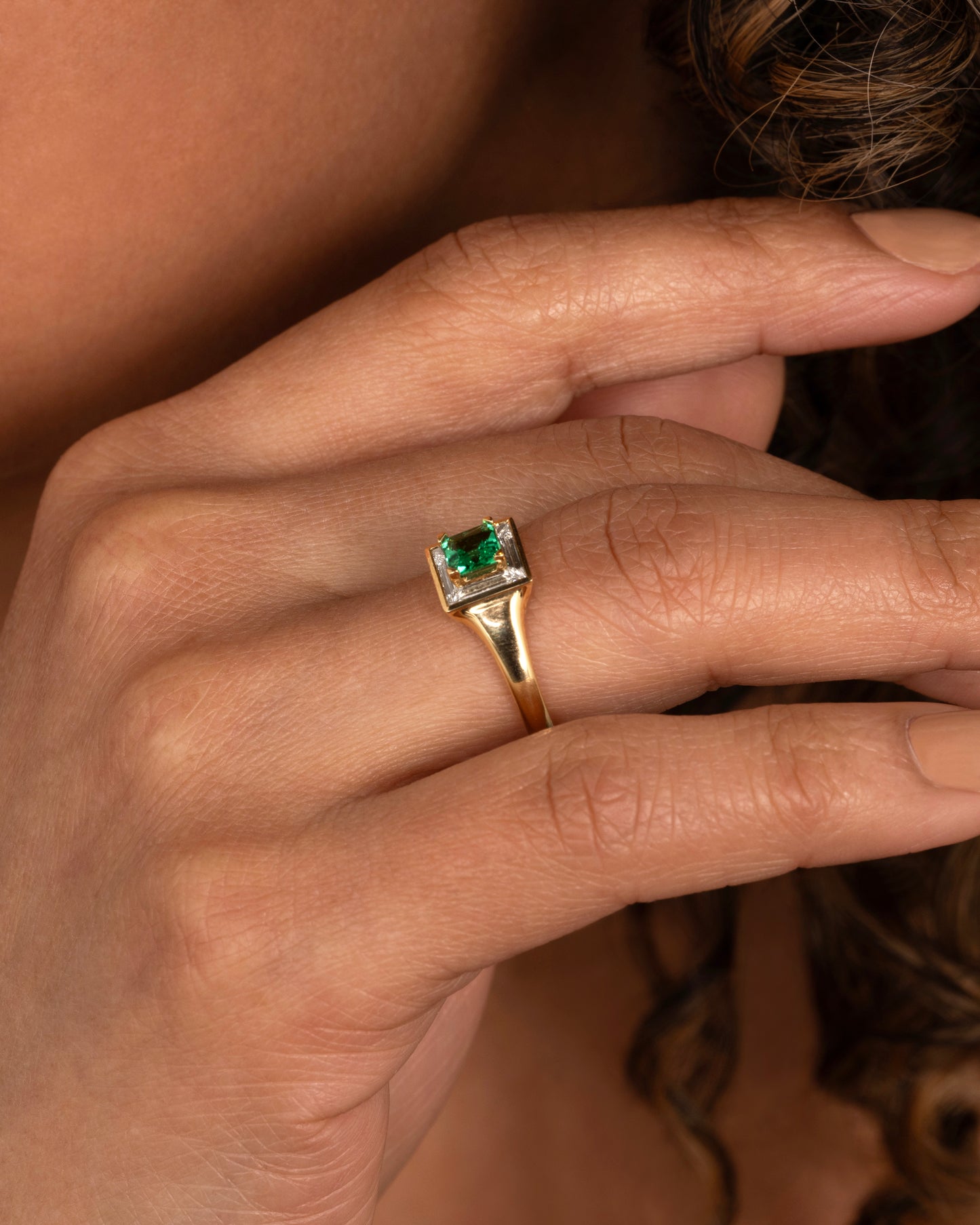 A yellow gold ring with a square emerald at the center surrounded by white baguette diamonds. Shown worn on a hand.