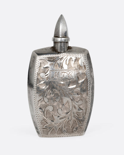A silver perfume bottle with botanical etchings, a twist- off wand, and the engraved initials M.T.C. Shown from the front.