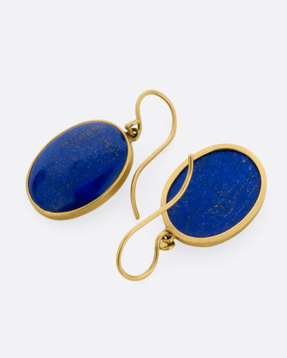 A pair of oval lapis earrings with yellow gold bezels and ear hooks. One earring is shown from the front, the other is shown from the back.