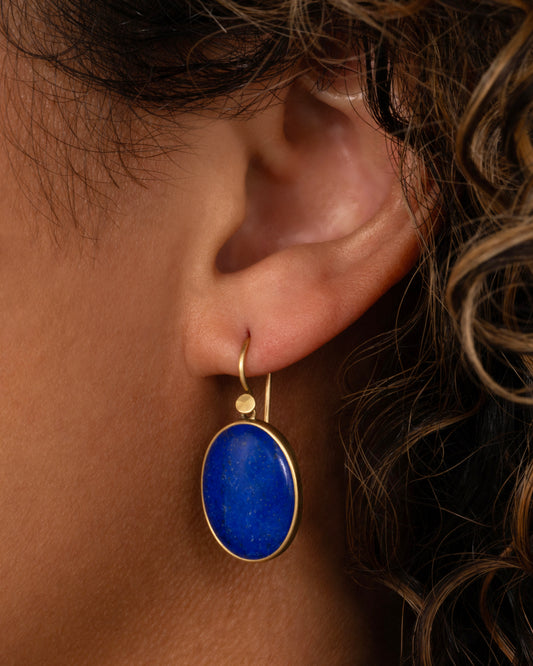 A pair of oval lapis earrings with yellow gold bezels and ear hooks. Shown from the front on a person.
