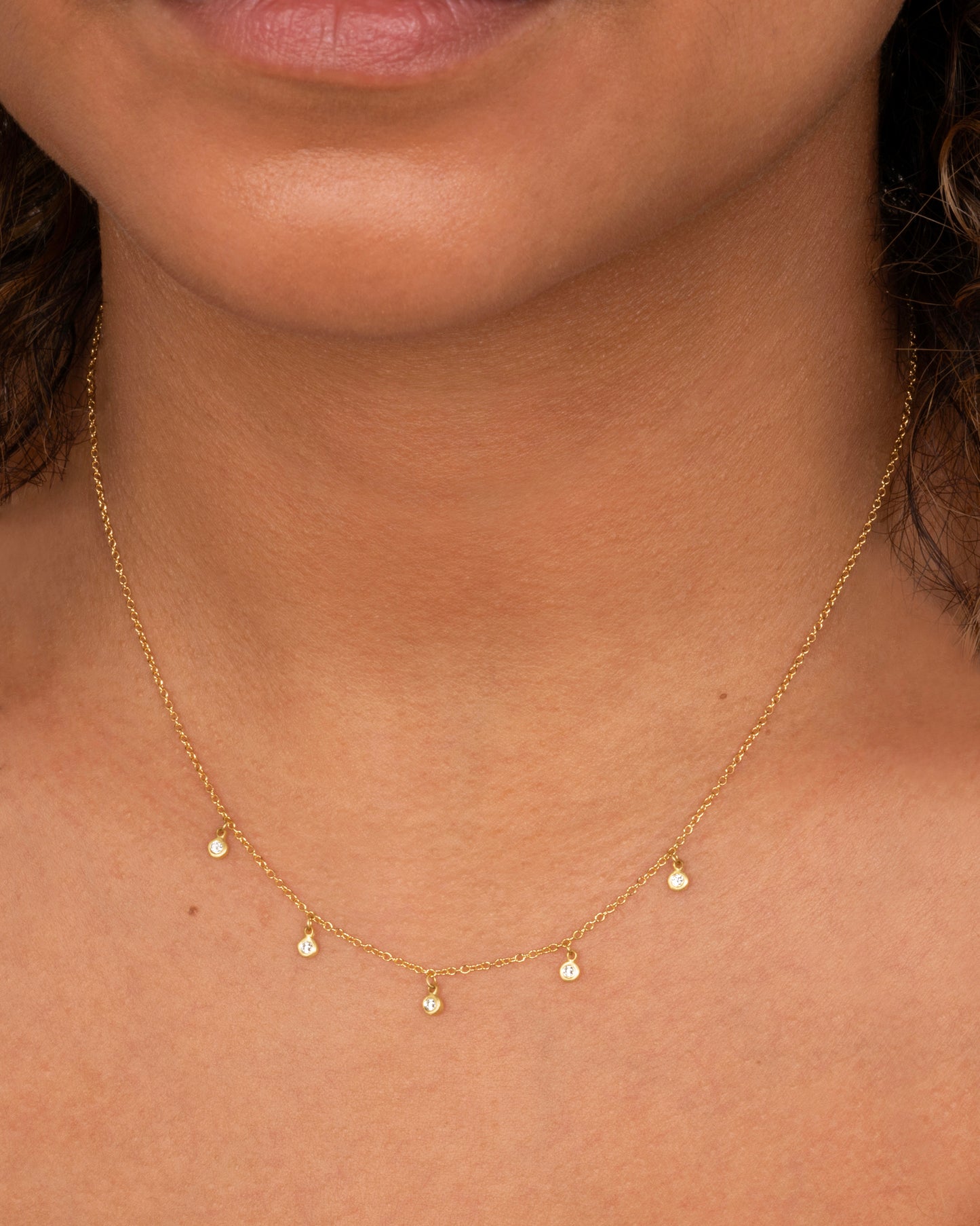 A yellow gold necklace with five brilliant bezel set diamond charms on a cable chain. Shown worn around the neck.