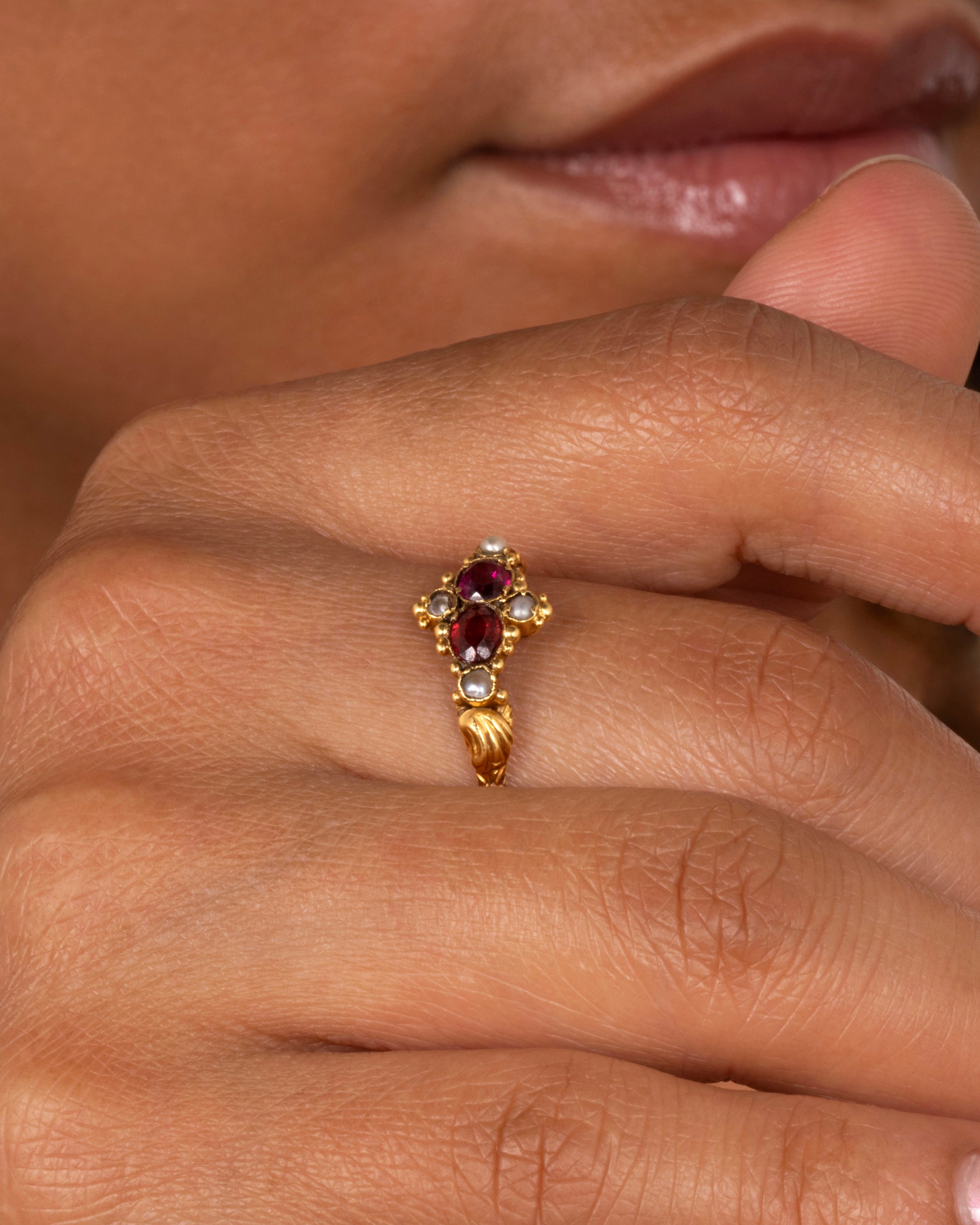 A yellow gold ring with garnets and seed pearls. Shown on a finger.