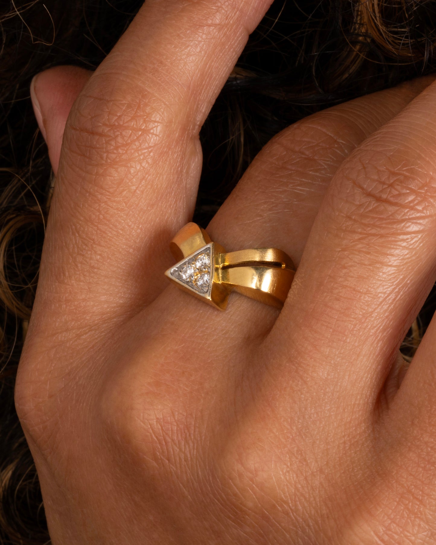 A yellow gold ring with a curvy band and three diamonds set in a triangular shaped setting. Shown worn on a finger.