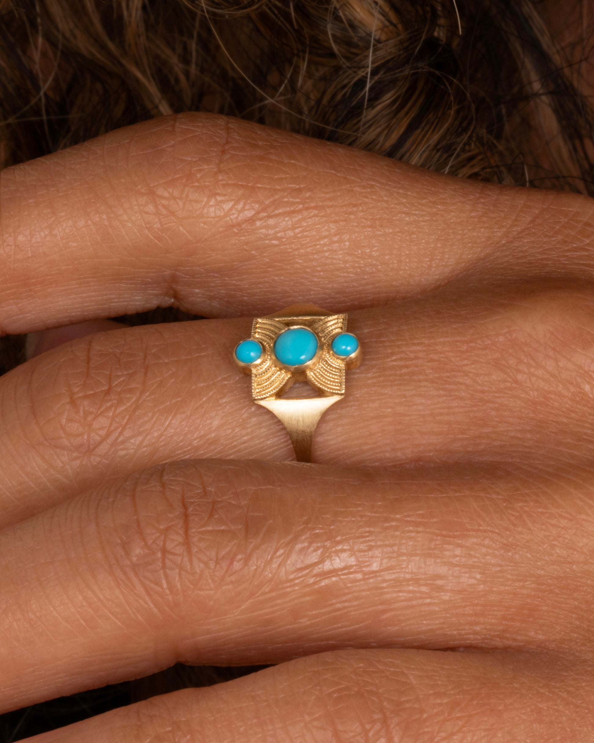 A yellow gold hourglass shaped ring with three stacked bezel set turquoises. Shown worn on a finger.
