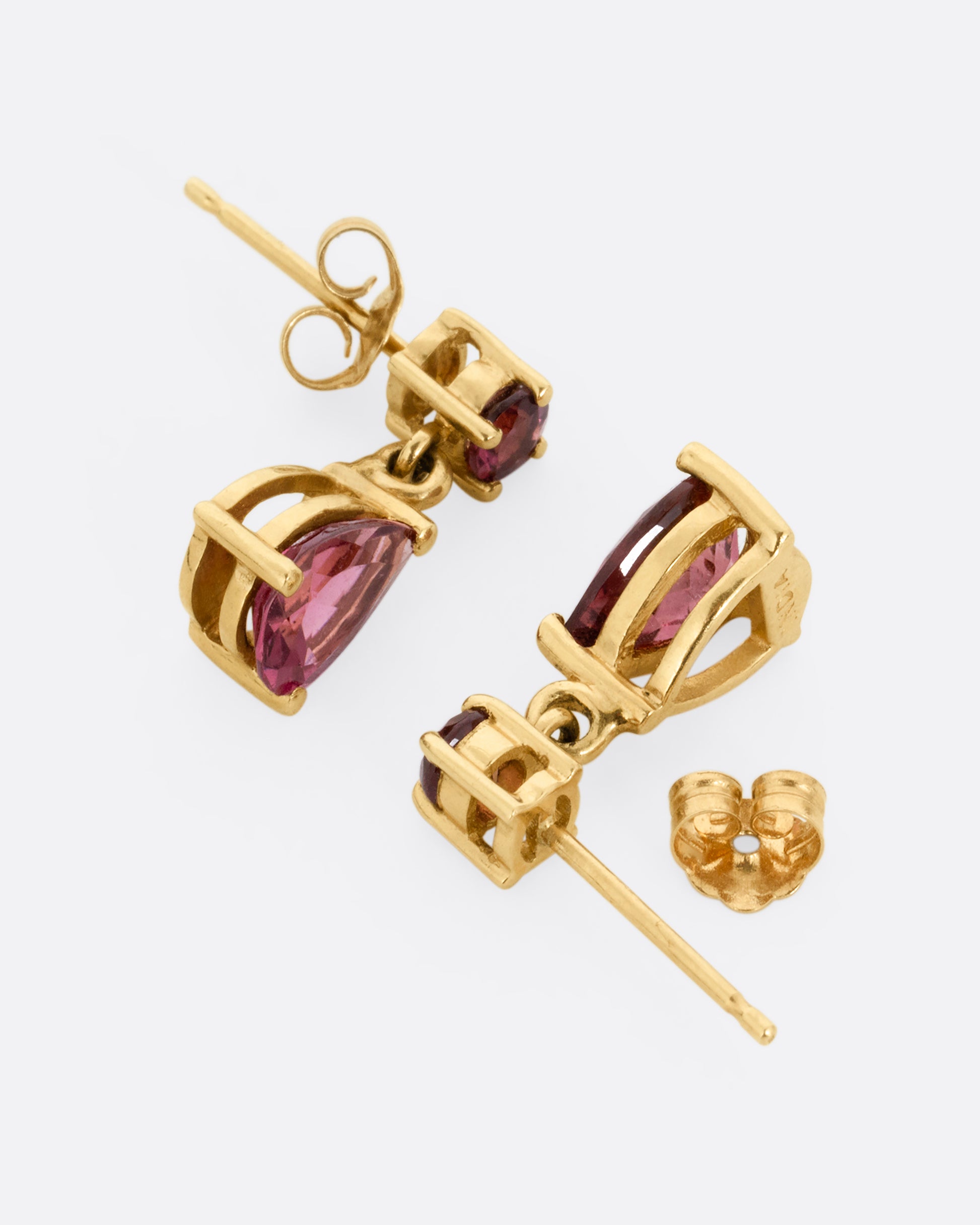 Yellow gold earrings with round prong set pink tourmalines at the top and teardrop shaped pink tourmalines at the bottom. One earring is shown lying on its side from the bottom, the other is shown open on its side from the top. 