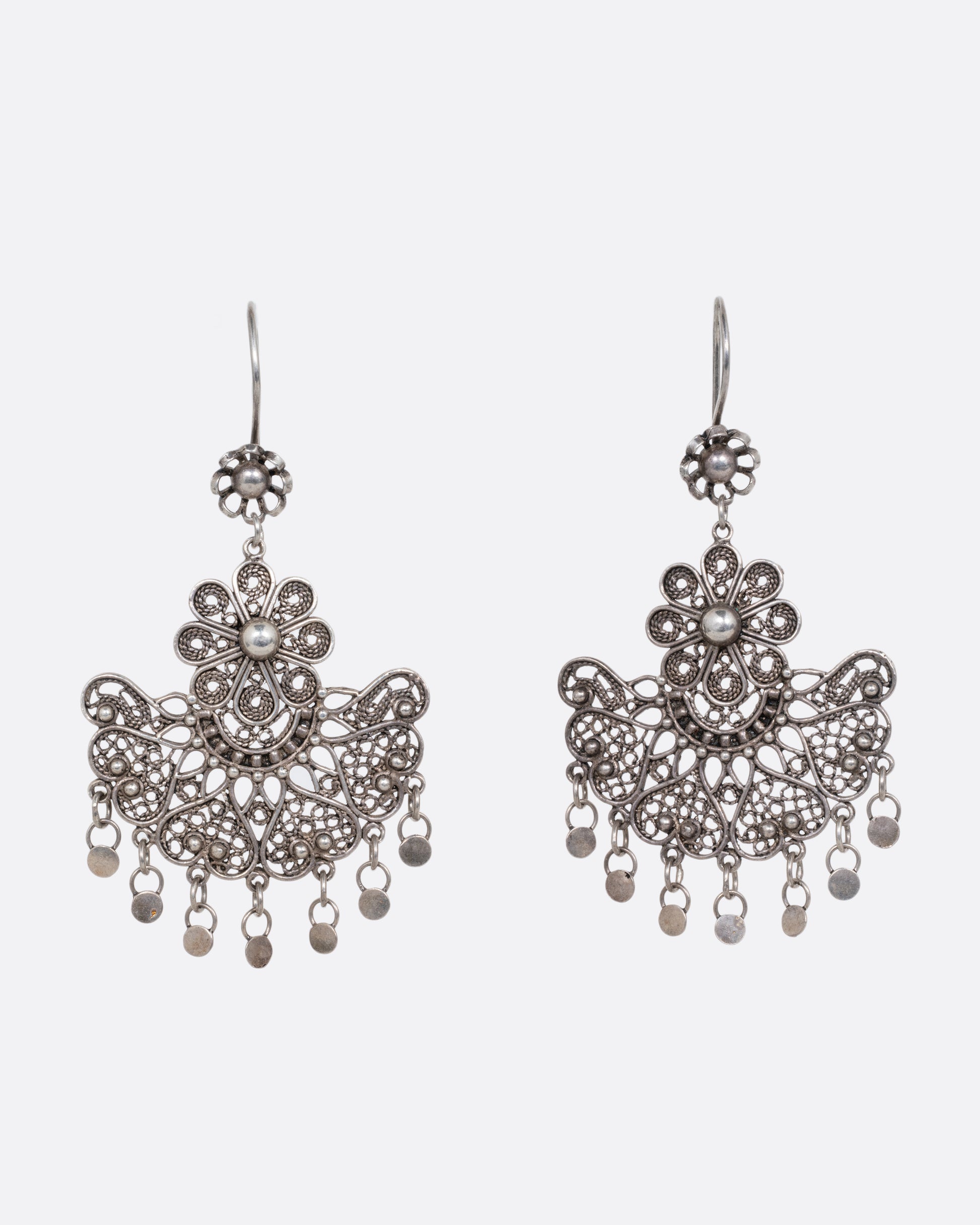 A pair of silver chandelier earrings with daisies on the ear hooks and filigree work below. There are seven tiny dangling disc charms hanging at the bottom of each earring. Shown from the front.