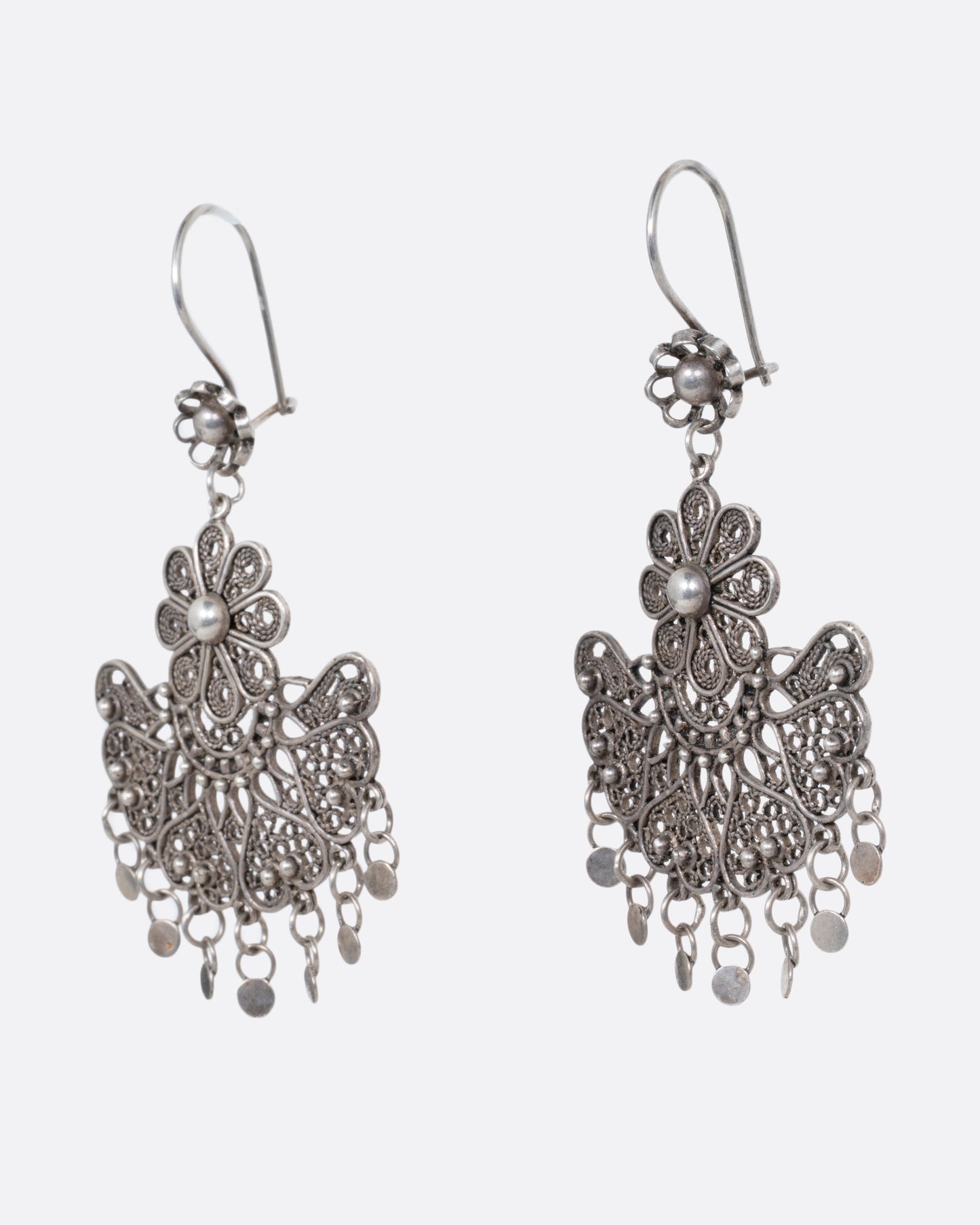 A pair of silver chandelier earrings with daisies on the ear hooks and filigree work below. There are seven tiny dangling disc charms hanging at the bottom of each earring. Shown from the side.