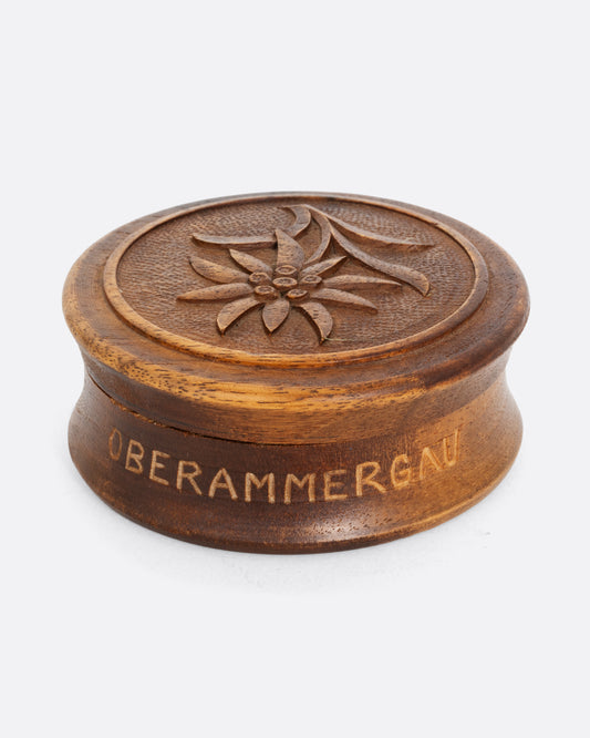 A circular hand carved wooden box with 'Oberammergau', a town in Germany, around its base and a flower on its top. Shown from the side.